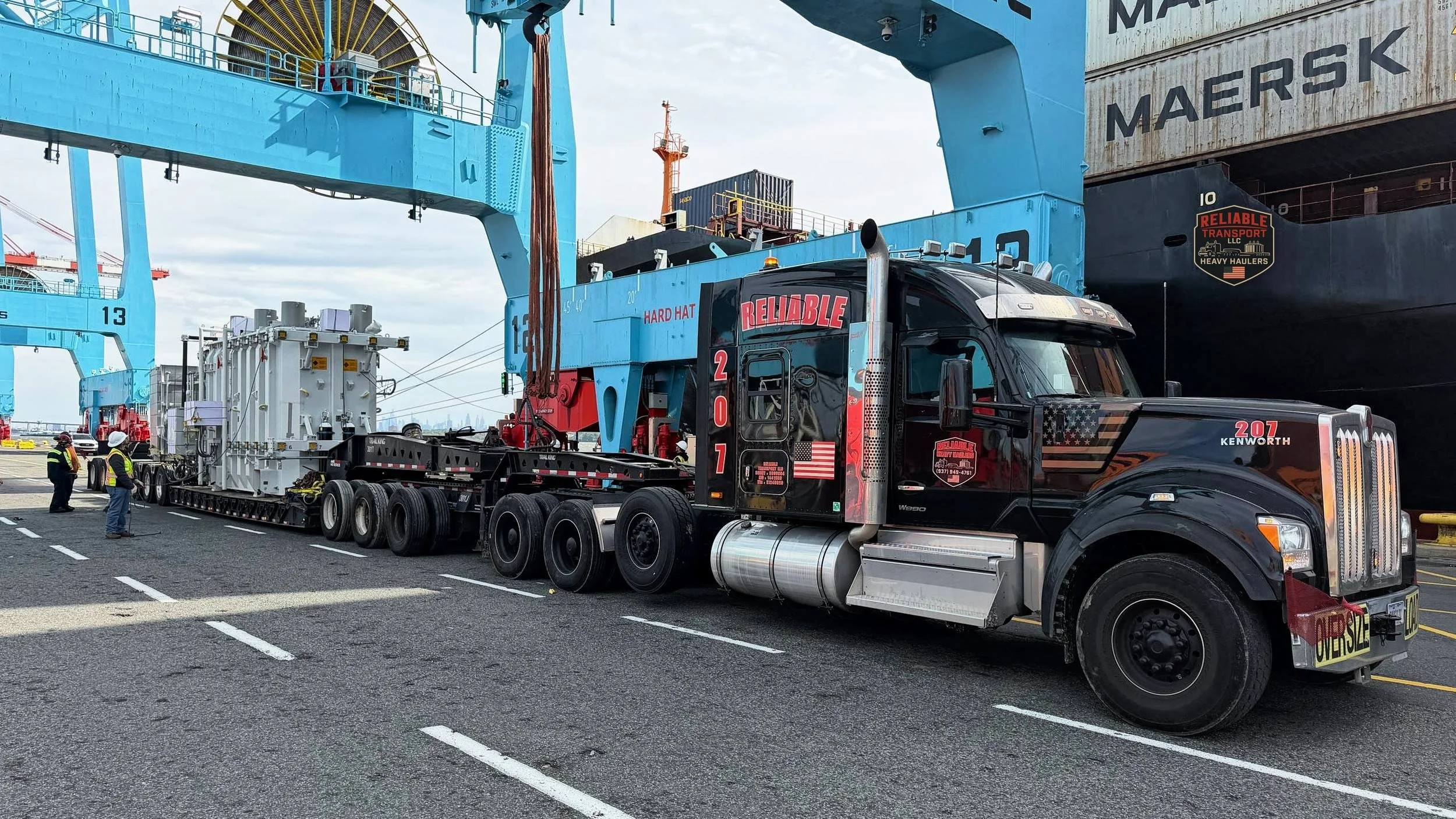 Black heavy haul tractor pulling a large transformer on a multi axle trailer beneath tall blue port cranes