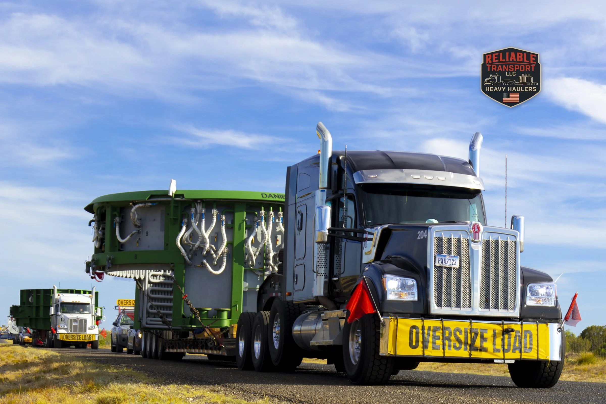 Black heavy haul truck with an OVERSIZE LOAD banner pulling a wide green load on a multi axle trailer along a two lane highway, with a second wide load and escort vehicles behind