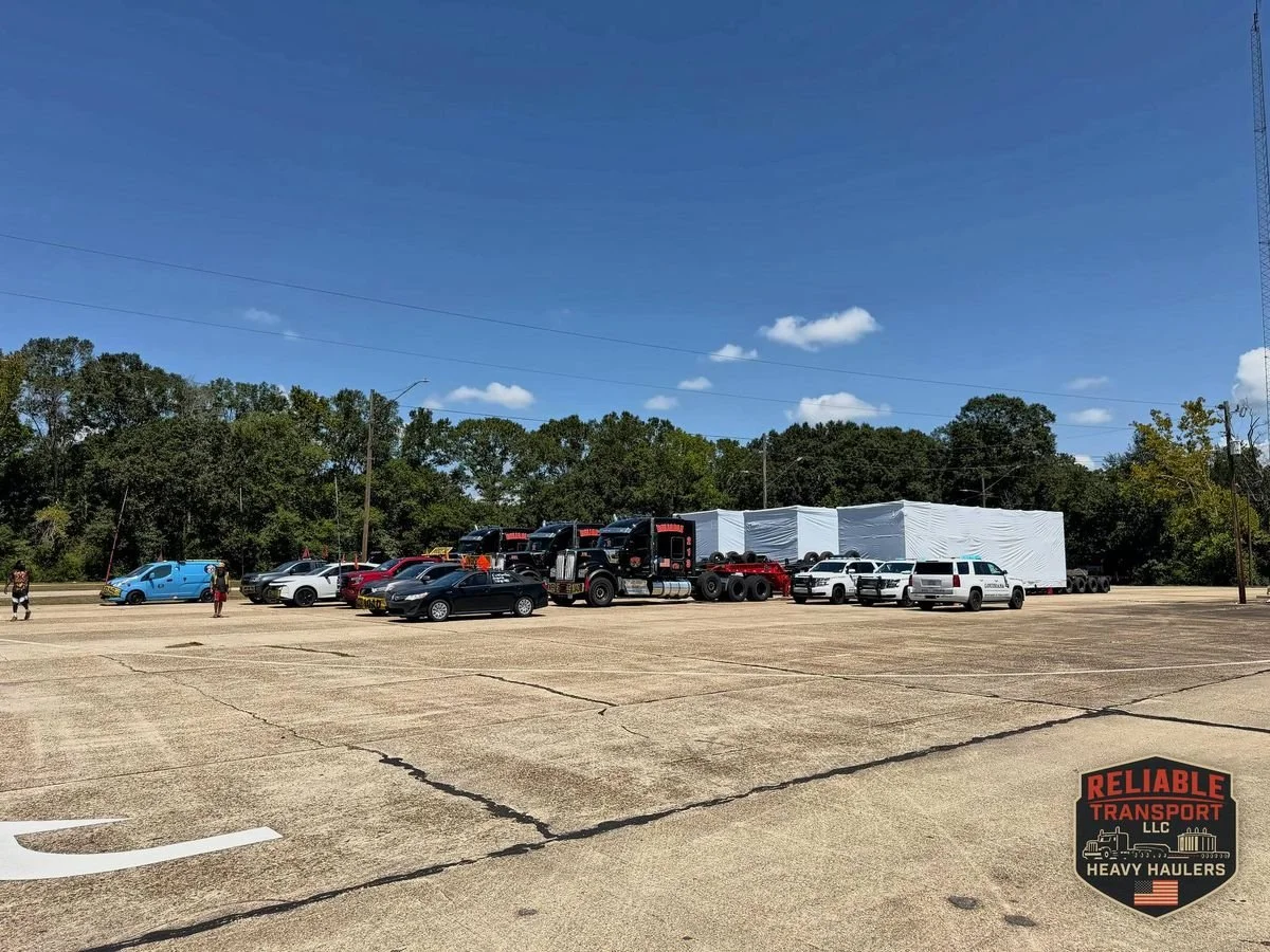 Escort vehicles and black trucks lined up in a paved staging lot, with white wrapped loads on trailers behind