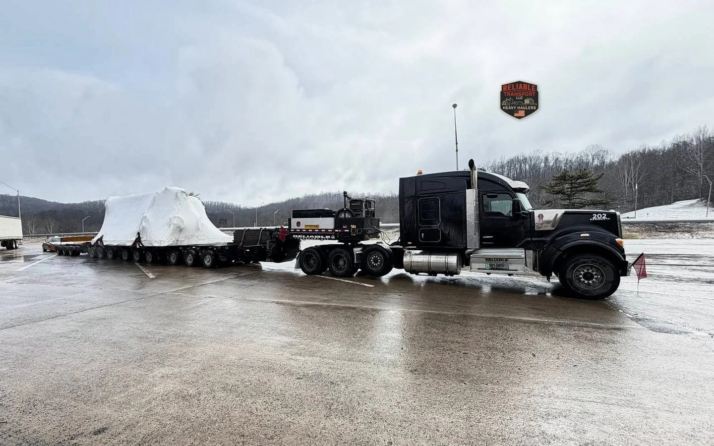 Black heavy haul tractor pulling a white wrapped load on a long multi axle trailer under a cloudy sky
