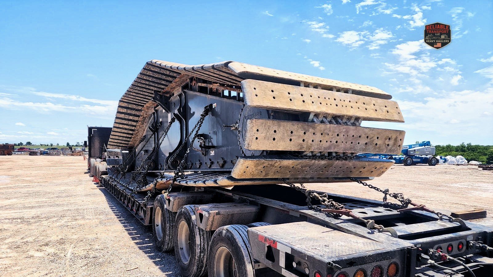 Large crawler track pad assembly chained to a multi axle trailer in a gravel yard under a blue sky