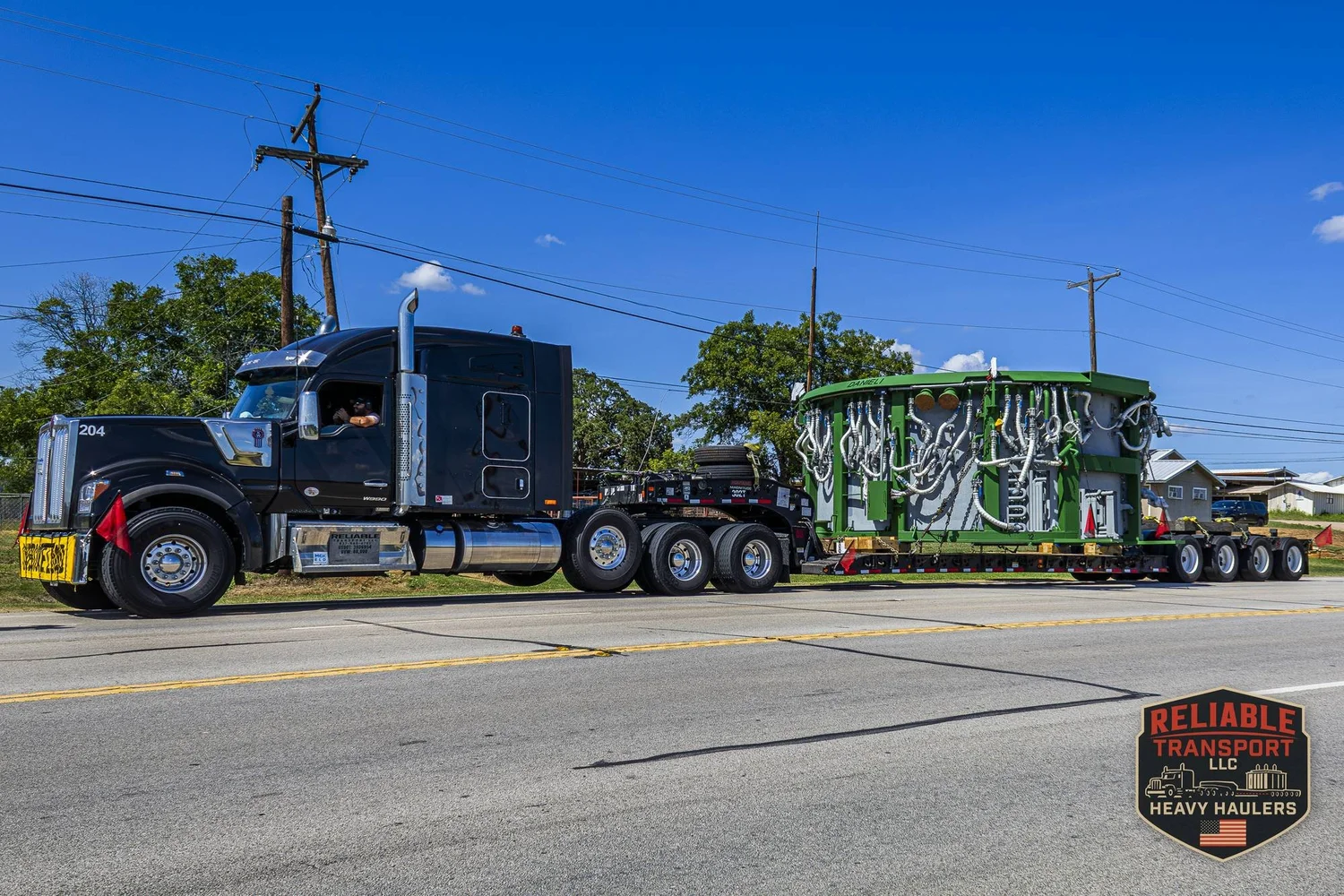 Black heavy haul truck pulling a wide green load on a multi axle trailer along a city street under power lines