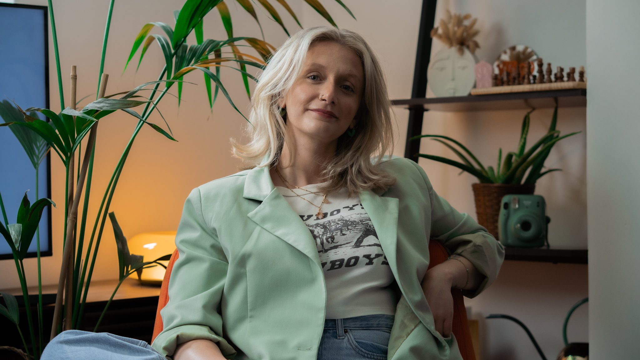 A woman with shoulder-length blonde hair, wearing a light green blazer over a graphic t-shirt, sits in a cozy room surrounded by houseplants, with a warm light illuminating the background.