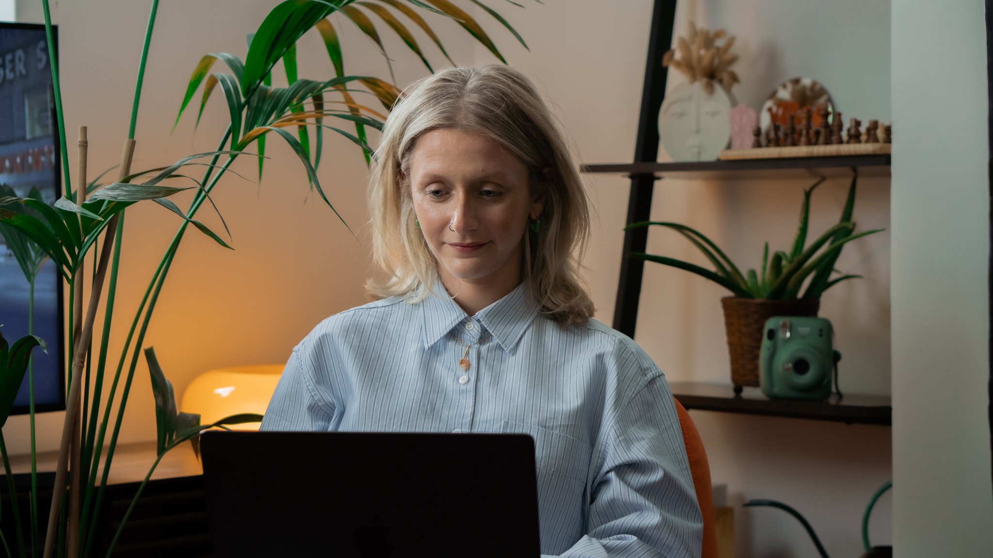 A young woman with blonde hair, wearing a light blue striped shirt, sits at a desk looking at a laptop screen in a cozy room with warm lighting, green plants, shelves with decorative items, a chess set, and a green speaker.