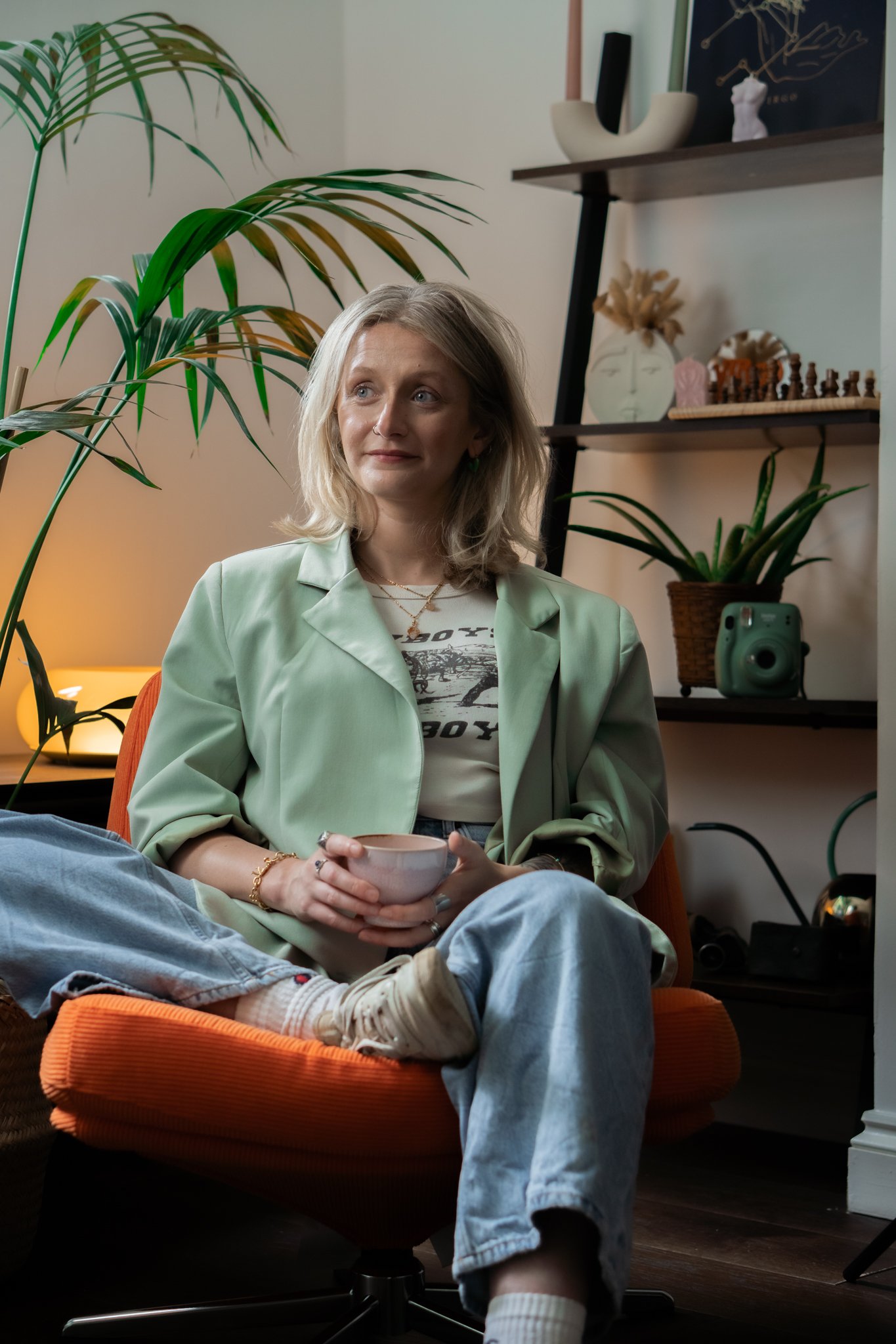 A woman with blonde hair sitting on an orange chair, holding a white mug, wearing a light green blazer, white graphic T-shirt, and light blue jeans, in a cozy living room with plants and decorative items.