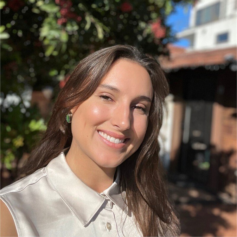 A young woman with long brown hair smiling outdoors on a sunny day, wearing a light-colored sleeveless blouse and earrings, with trees and a building in the background.