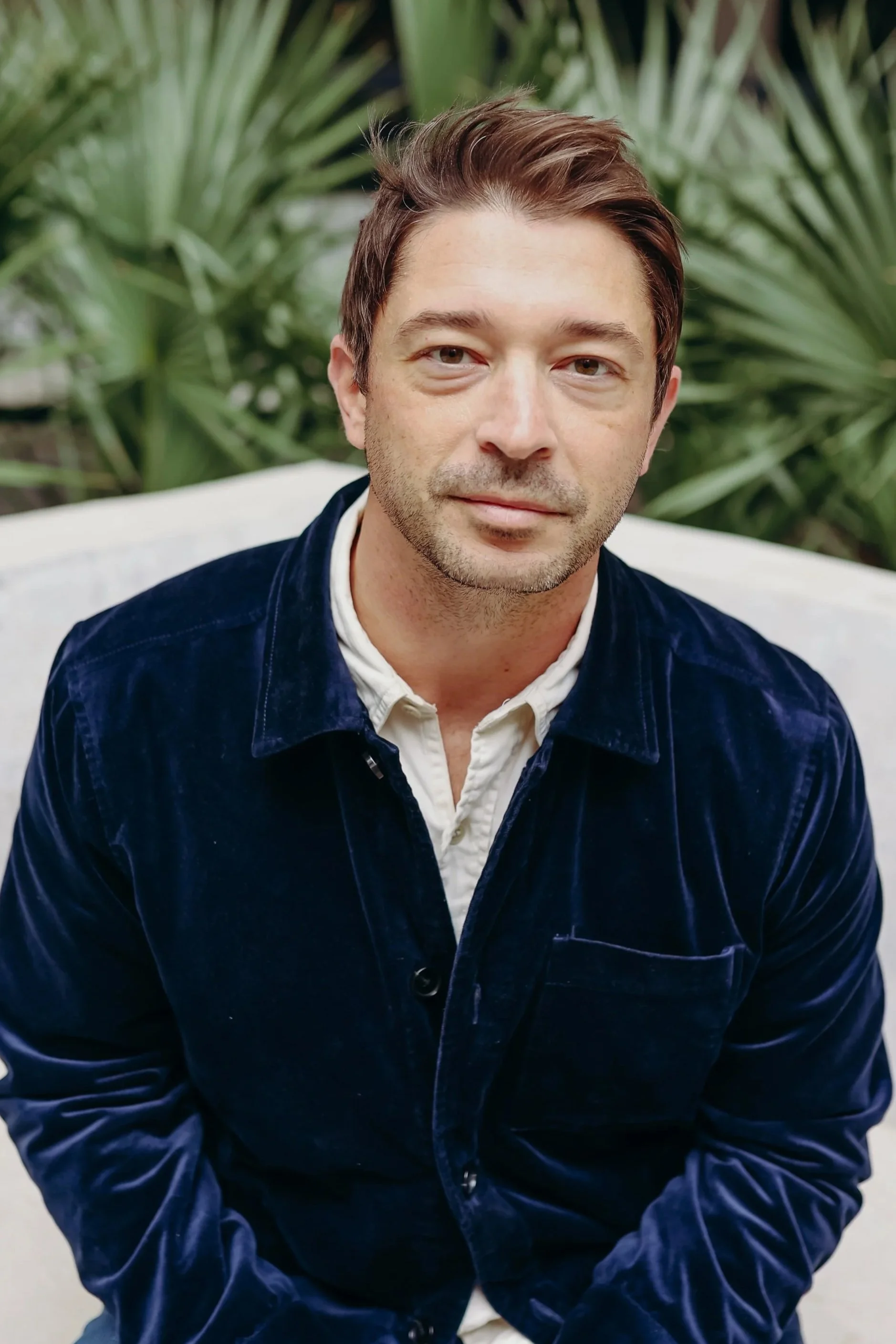A man with brown hair and a slight beard, wearing a dark blue velvet jacket over a white shirt, sitting on a white couch outdoors with green tropical plants in the background.