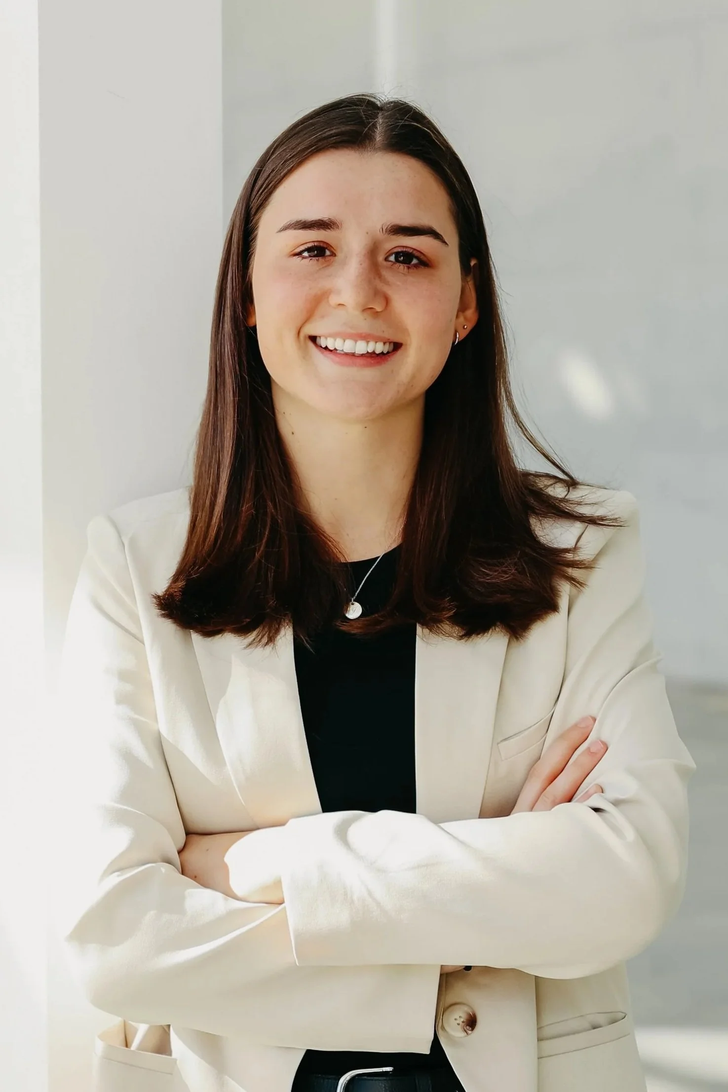 A young woman with long brown hair, wearing a cream blazer and black top, smiling with arms crossed in a bright, neutral-colored room.