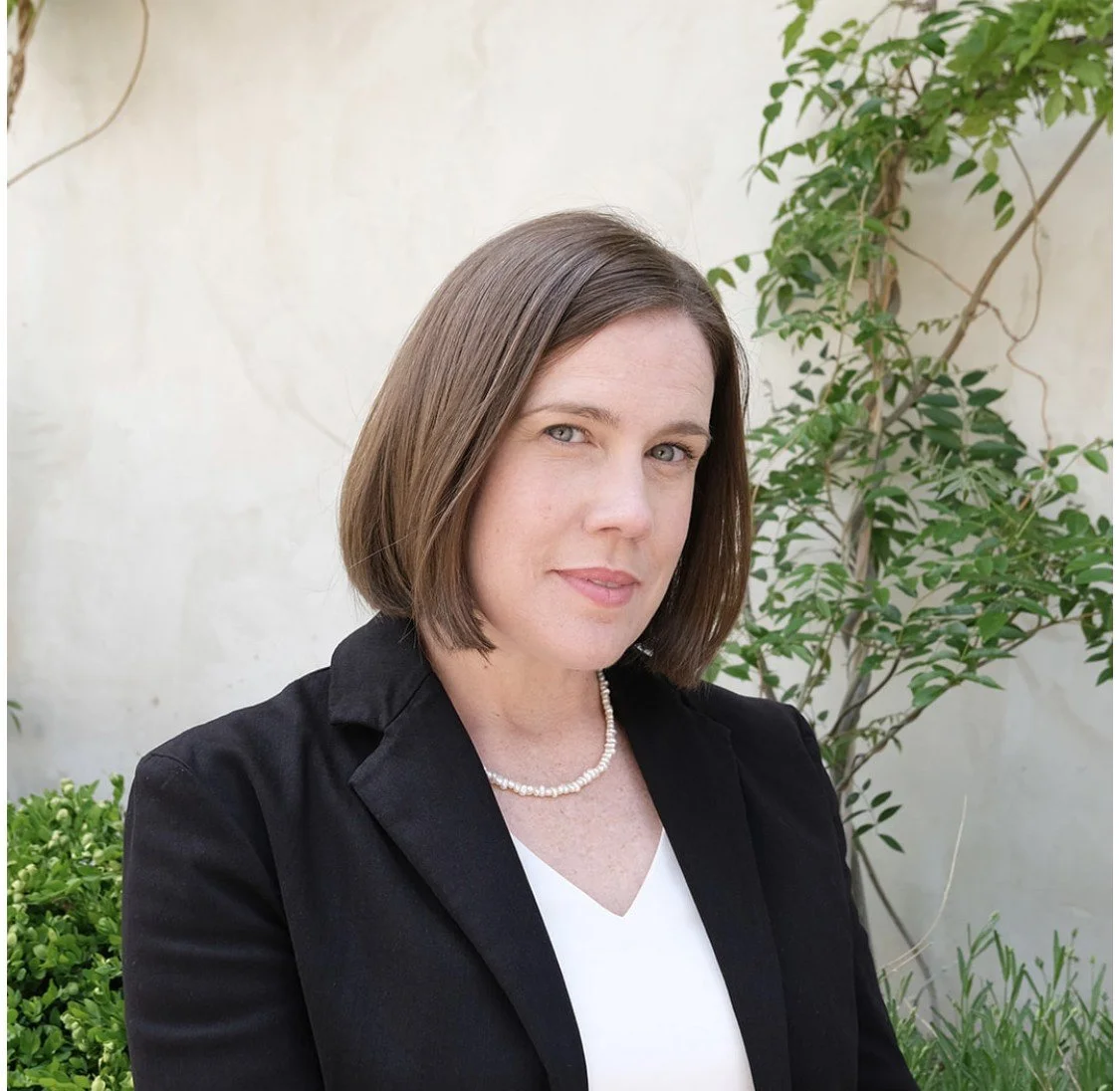 A woman with shoulder-length brown hair, wearing a black blazer, white top, and pearl necklace, posing outdoors with green plants and a light-colored wall in the background.
