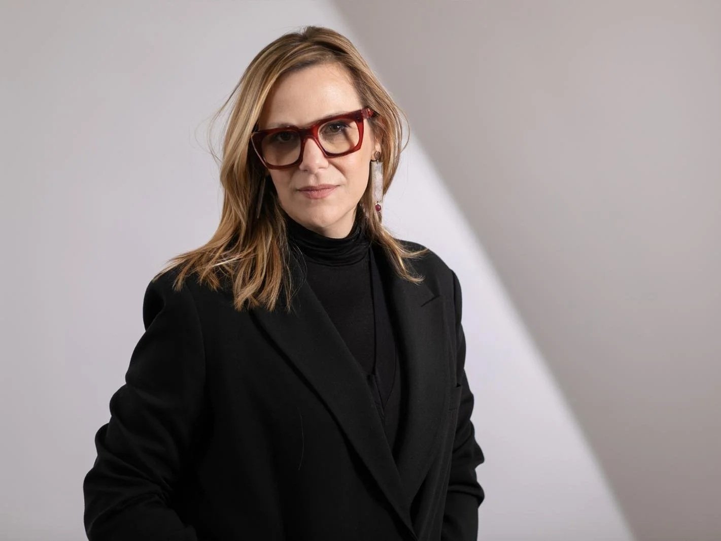 A woman with shoulder-length blonde hair, wearing red glasses, gold earrings, a black turtleneck, and a black blazer, stands in front of a light gray background.