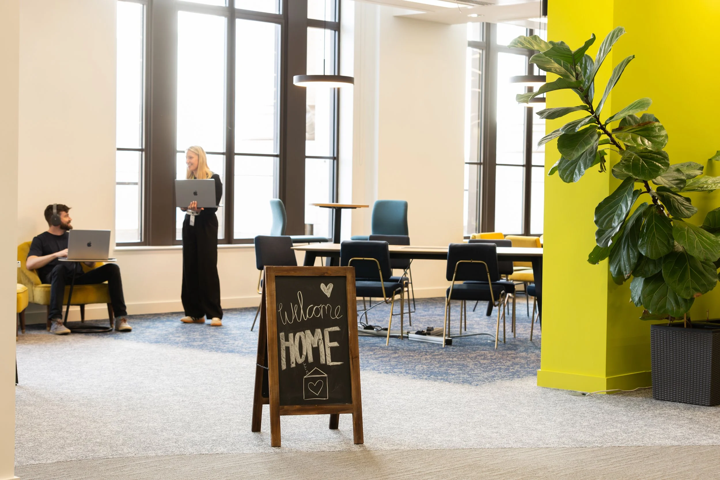 Modern office space with large windows, a yellow accent wall, and a potted plant. There are two people with laptops and headphones, one sitting and one standing, and a chalkboard sign that reads 'Welcome HOME' with a small heart and house drawing.