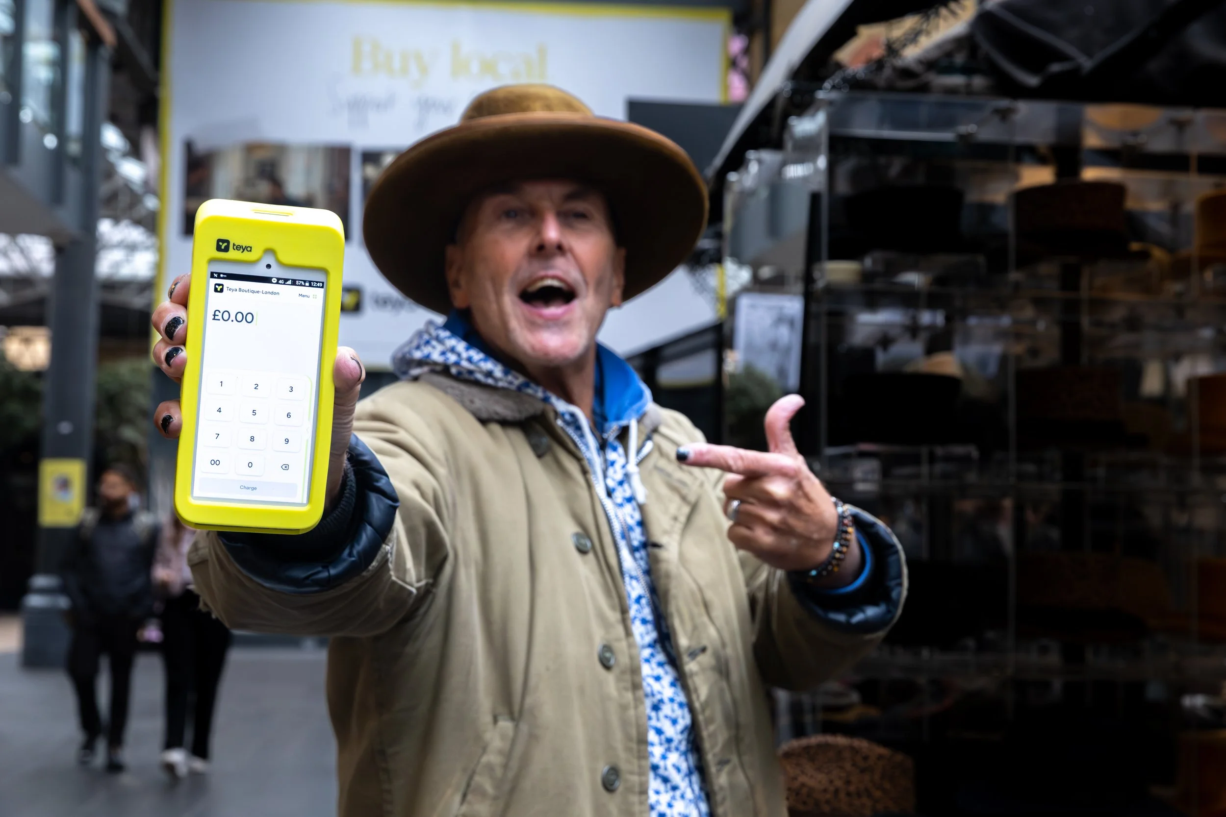 A man wearing a wide-brimmed hat and beige jacket holding a yellow mobile phone showing a payment app, standing outdoors in a shopping area with shelves of hats behind him and a sign in the background.