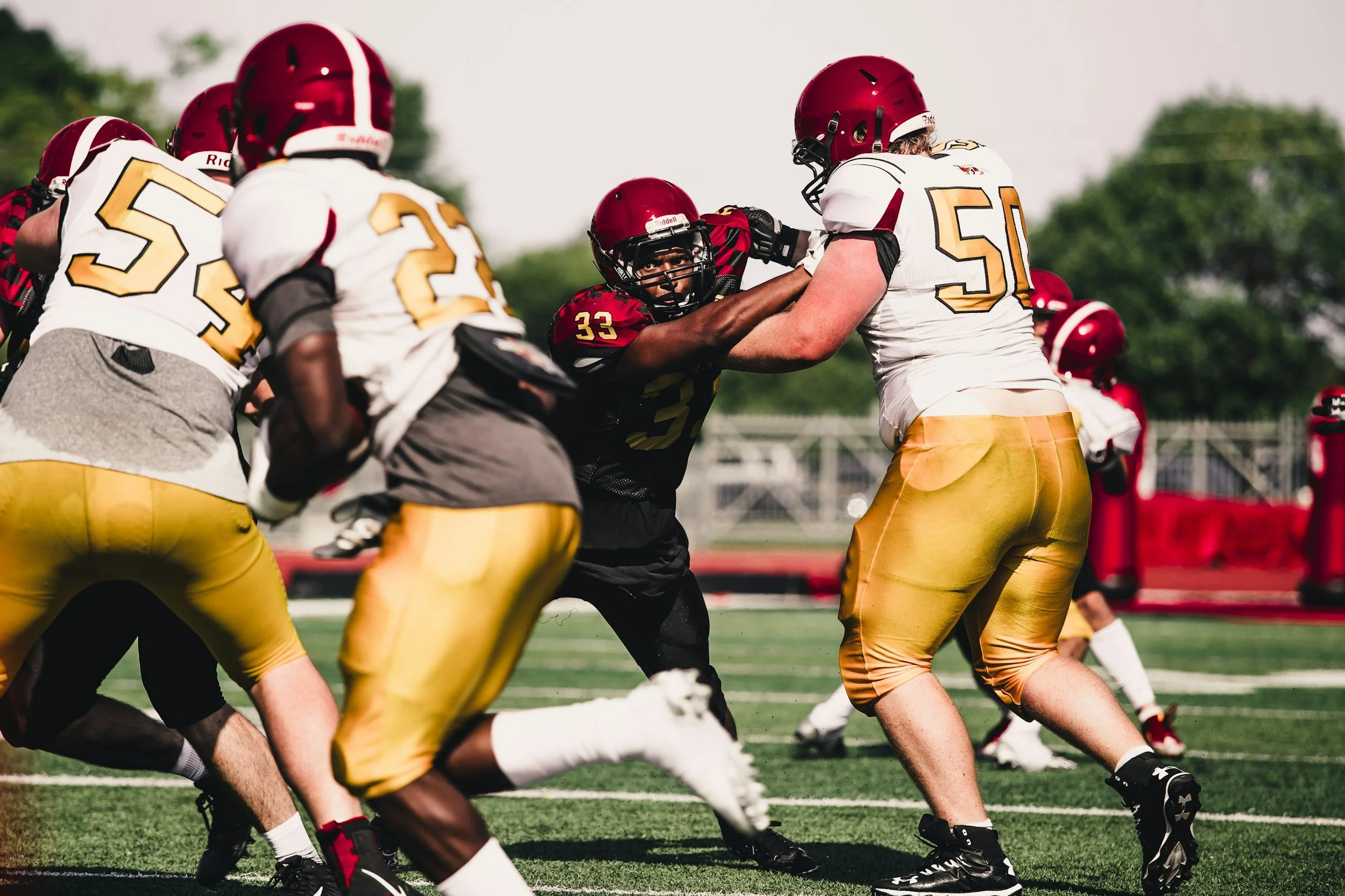 American football players during a game, with one player in black attempting to block a player in white and gold, while others prepare for the play.