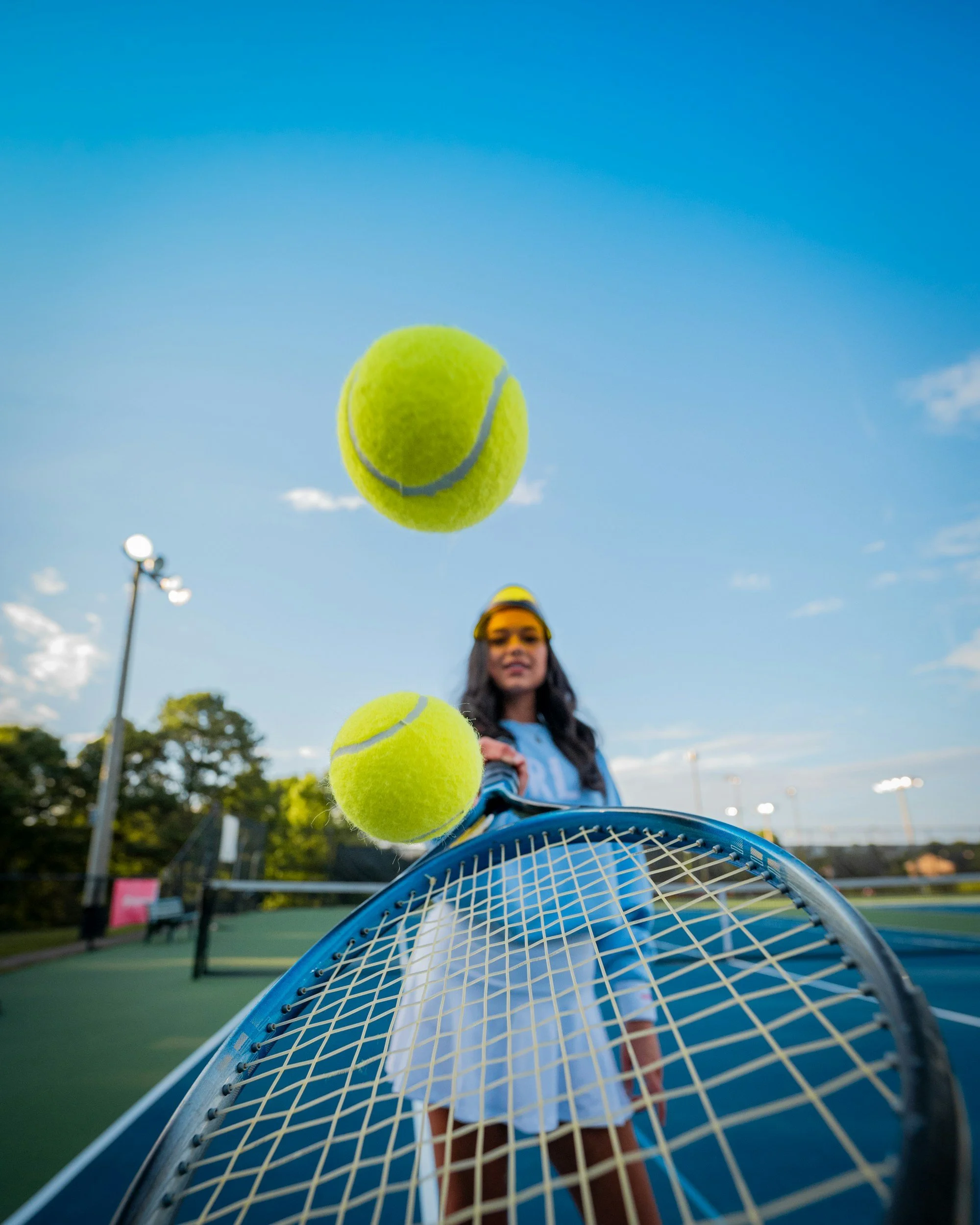 Teenage girl on tennis court with tennis balls in the foreground, blue sky, trees, and lamp posts in the background.