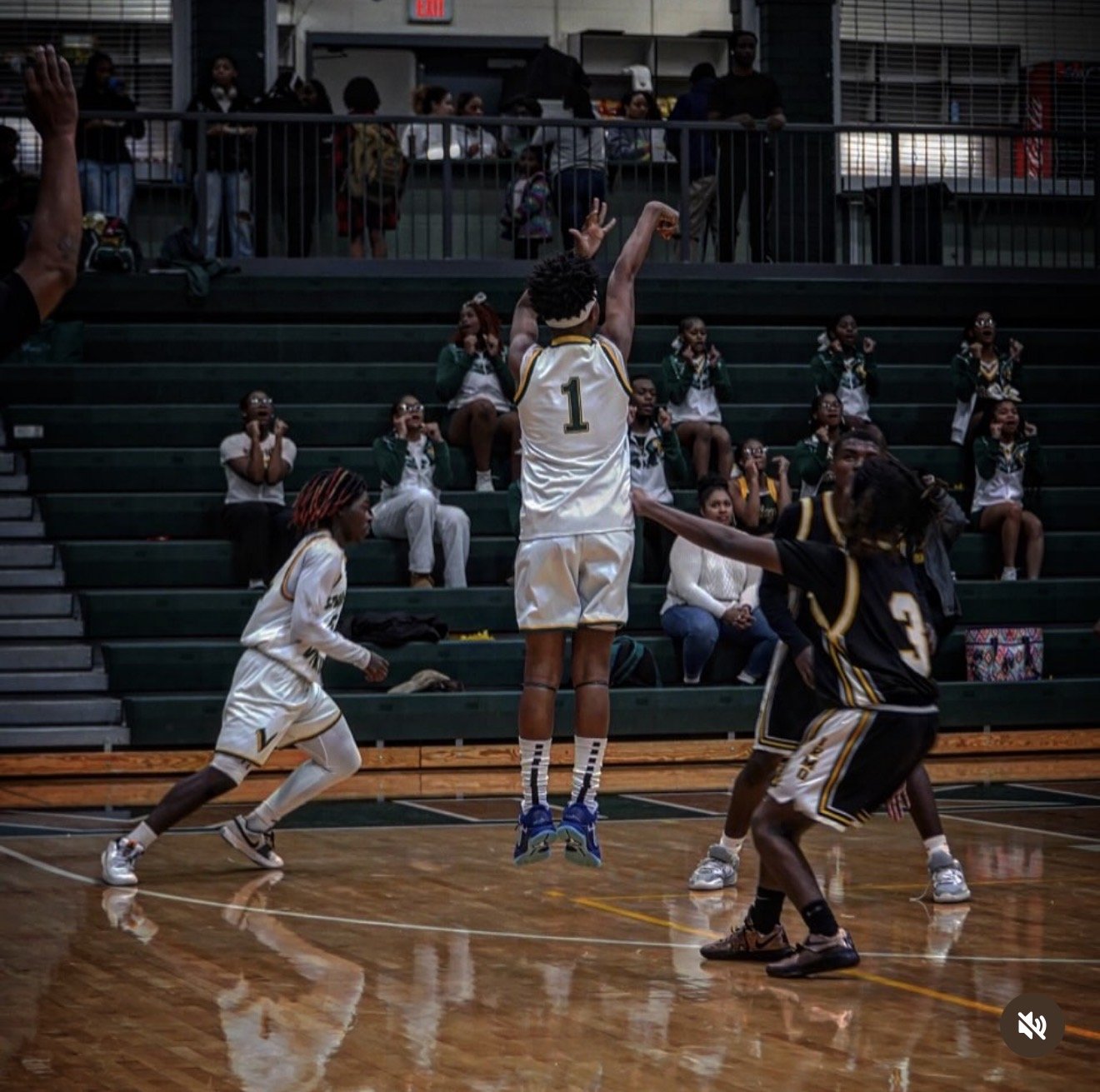A basketball game with a player wearing a white jersey with the number 1 jumping to shoot the ball, while another player in a black jersey with the number 3 defend. Spectators, including children in green and white uniforms, observe from the bleachers.