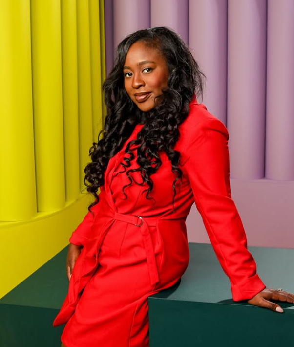 A woman with long curly black hair, wearing a red dress, sitting against a colorful background of yellow and purple vertical panels.