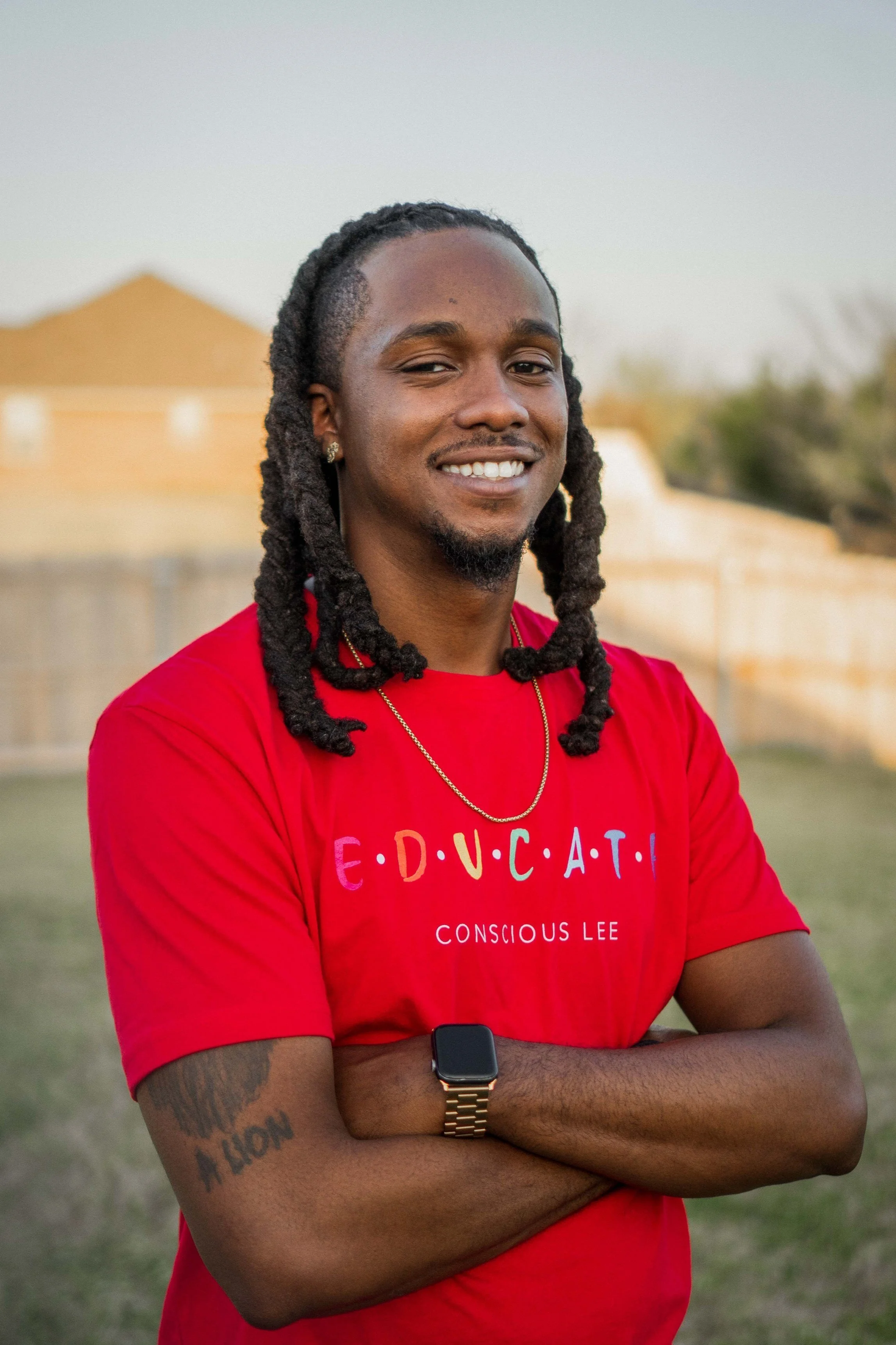 Man with long hair and beard smiling, wearing a red "EDUCATE Conscious Lee" t-shirt and a watch, standing outdoors with crossed arms.