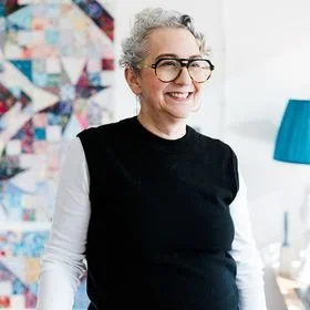 Smiling woman with curly gray hair and glasses, wearing a black and white top, standing in a bright room with colorful artwork and a blue lamp in the background.