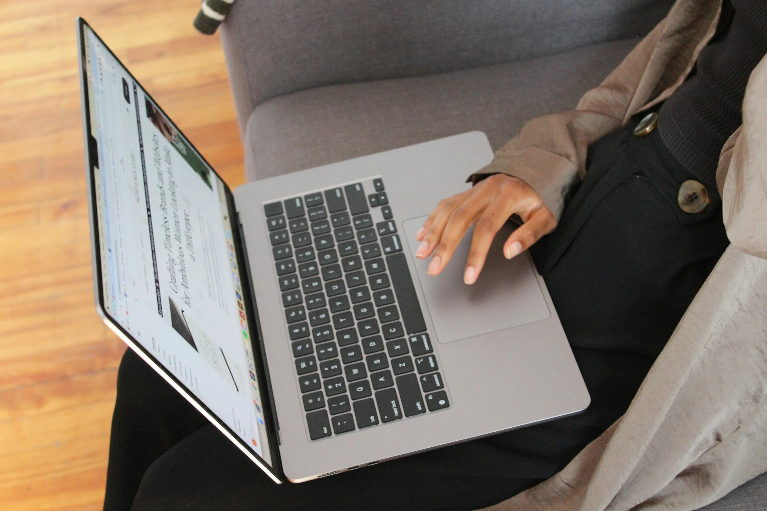 Person sitting on a gray chair using a silver MacBook laptop, with a wooden floor in the background.