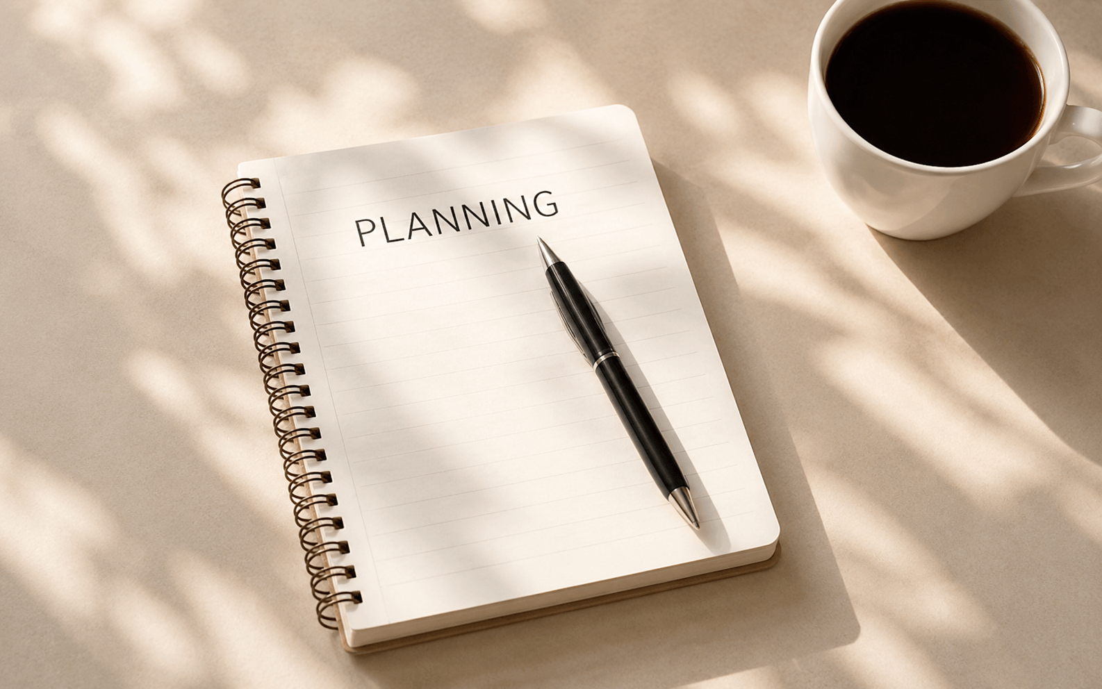 A spiral notebook with the word "PLANNING" written on the page, a black pen resting on it, and a white mug filled with black coffee on a beige surface with dappled sunlight.