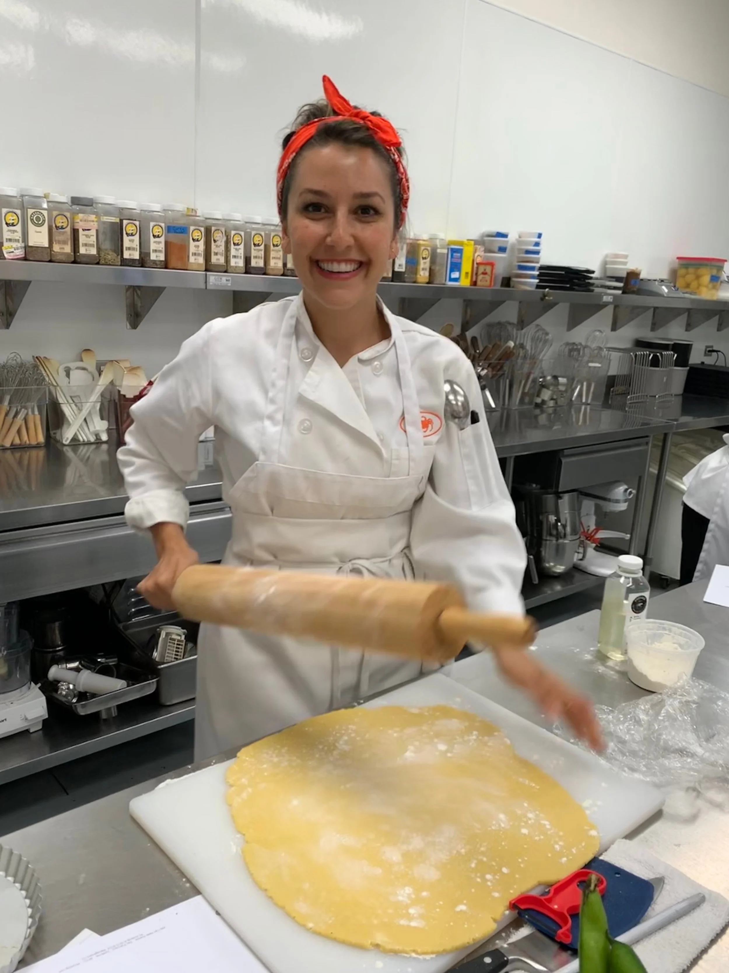 A baker in a white chef's coat and red bandana rolling out dough with a rolling pin in a commercial kitchen.