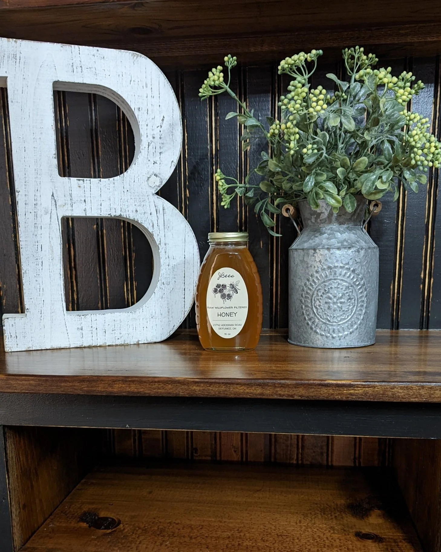 Decorative scene with a large white letter 'B', a jar of honey, and a gray metal vase with green foliage on a wooden shelf.