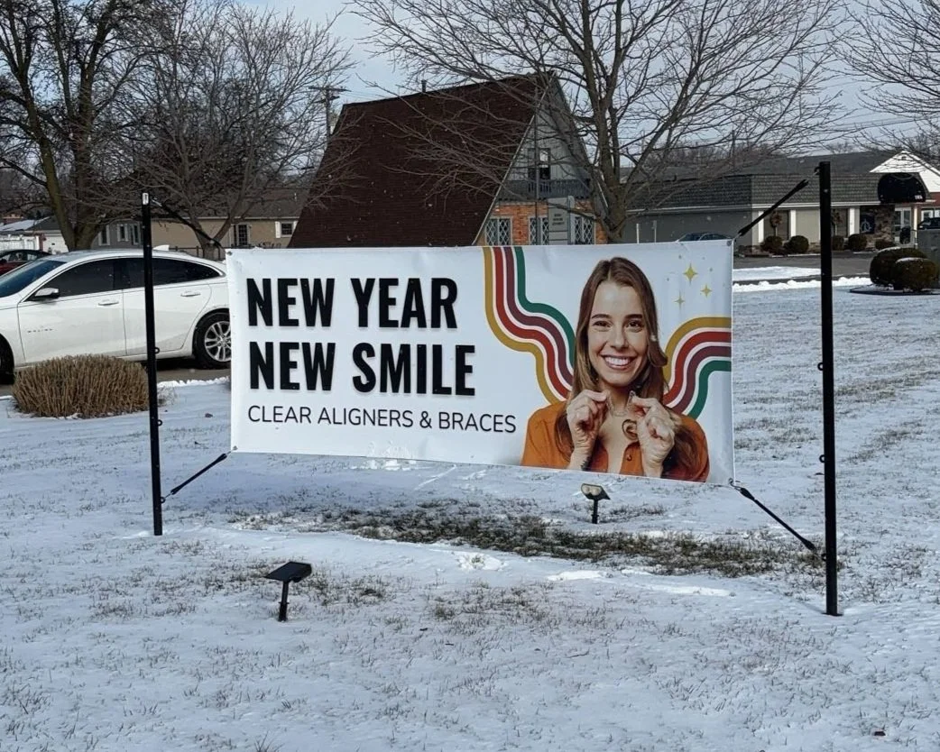 Outdoor advertisement banner for dental services, showing a smiling woman with braces, with text that reads 'New Year New Smile, Clear Aligners & Braces,' on a snow-covered lawn in a residential neighborhood.