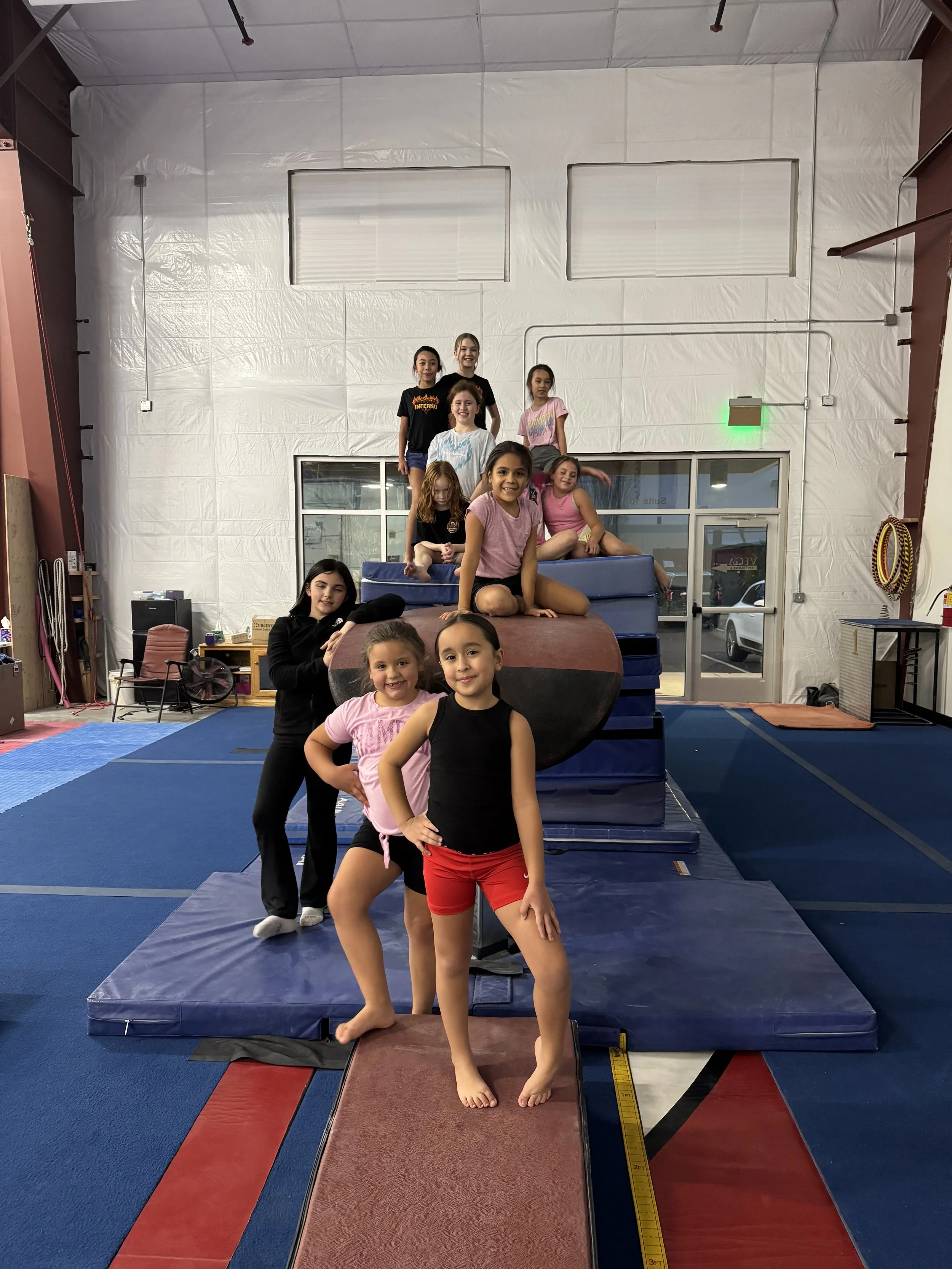 Group of children at a gymnastics gym, standing and sitting on a large foam climbing structure, smiling for the camera.
