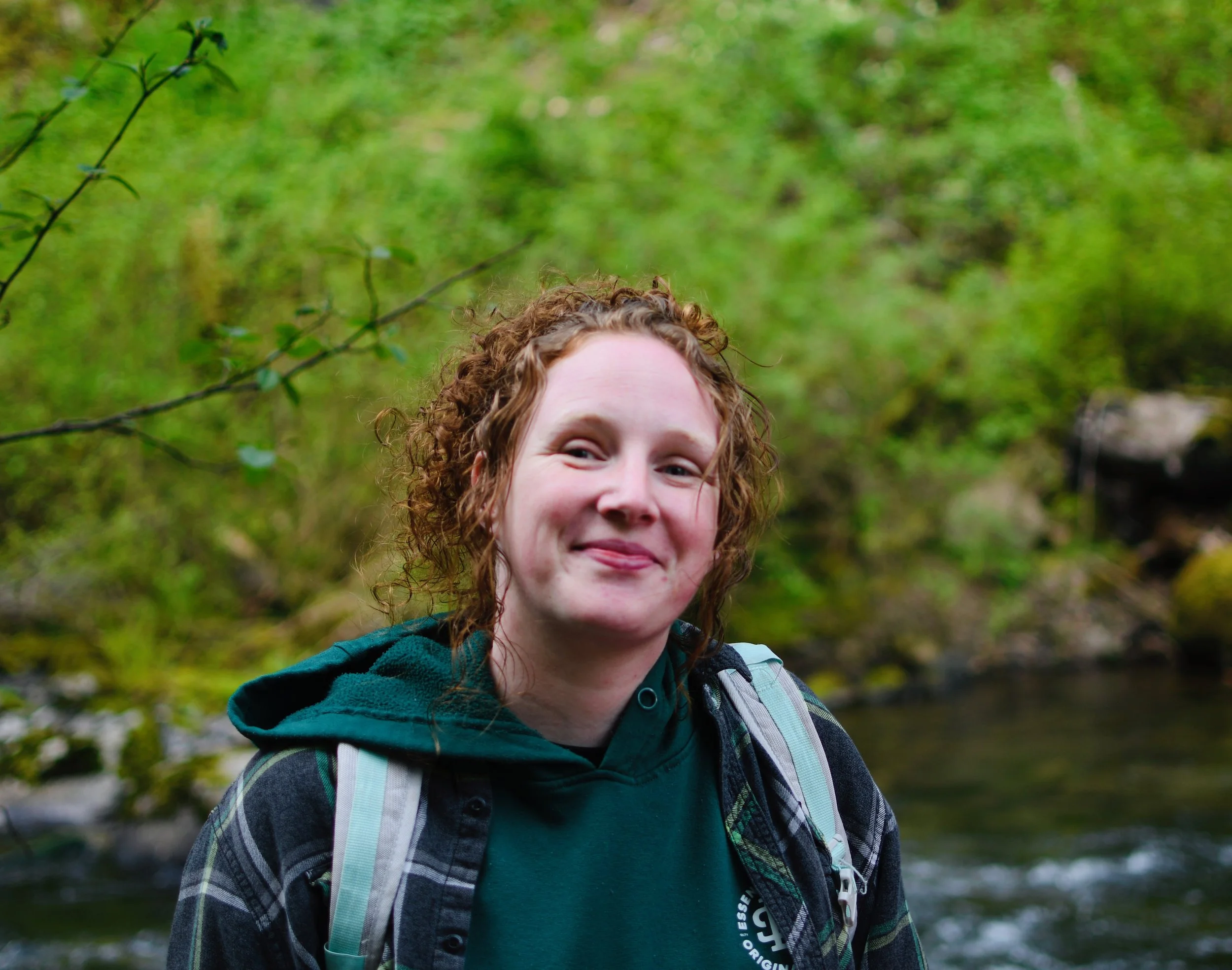 A young woman with curly red hair smiling outdoors near a creek, wearing a dark green hoodie and a plaid shirt with a backpack.