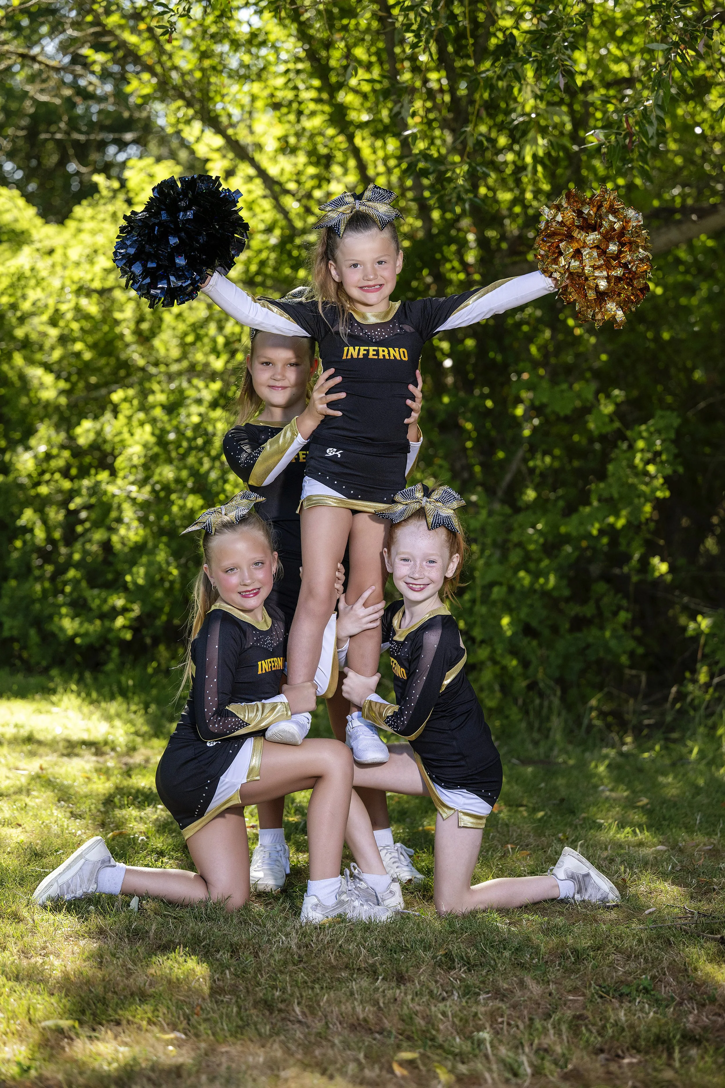 Four young cheerleaders in black and gold uniforms forming a pyramid outdoors, with green foliage in the background.