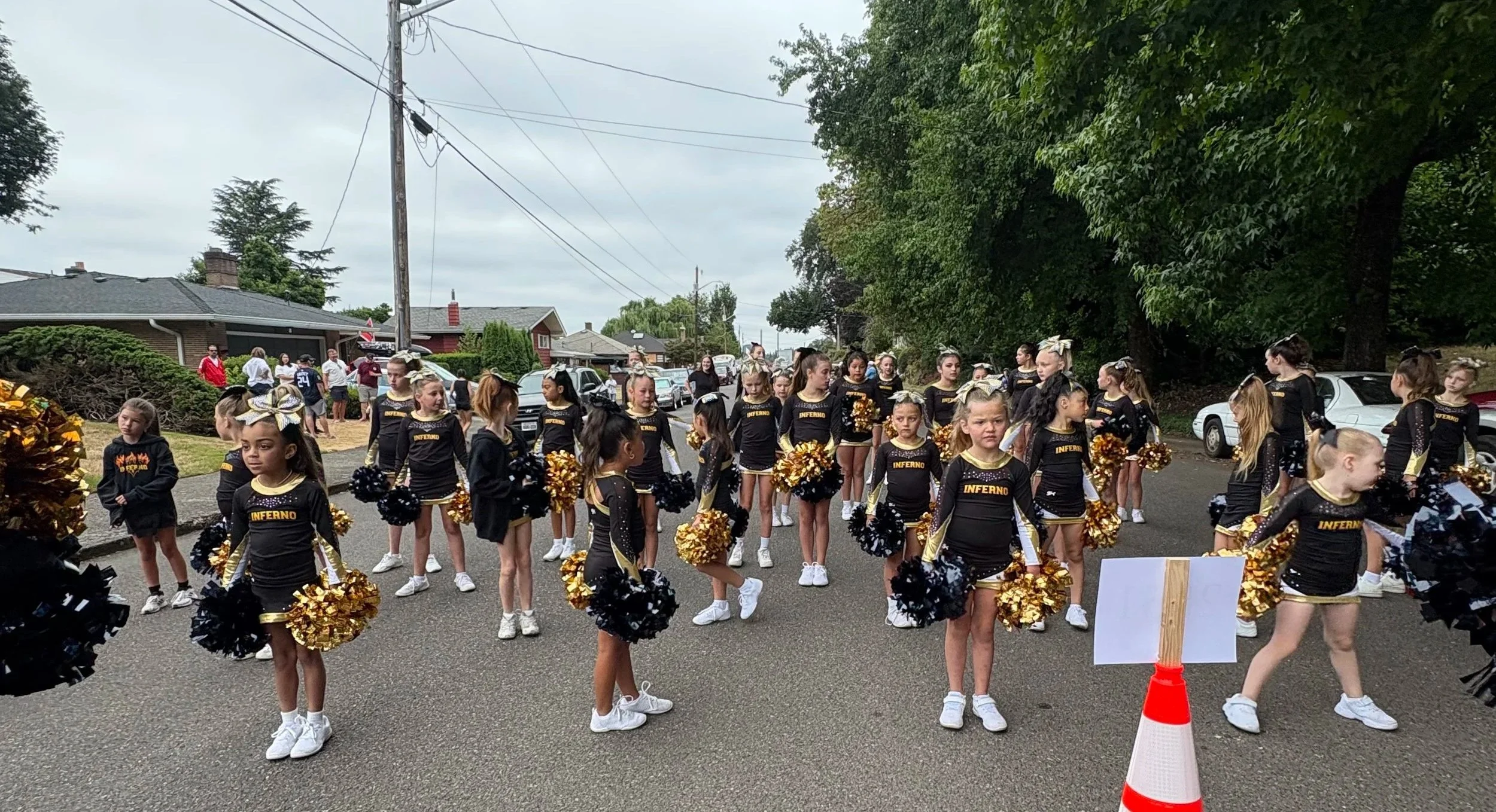 A group of young cheerleaders dressed in black and gold uniforms with pom-poms, preparing for a parade or performance on a residential street with houses, cars, and trees in the background.