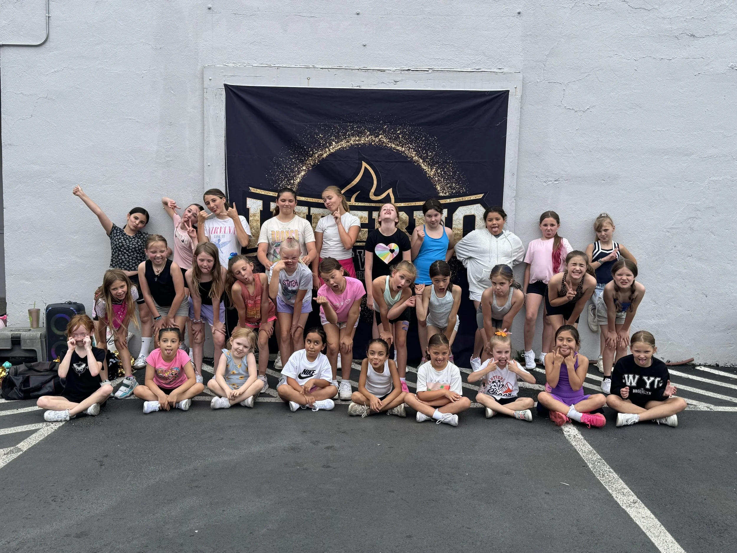 Group of young girls, some sitting and some standing, posing and making playful gestures in front of a backdrop with a heated logo. The setting appears to be outdoors on a parking lot.