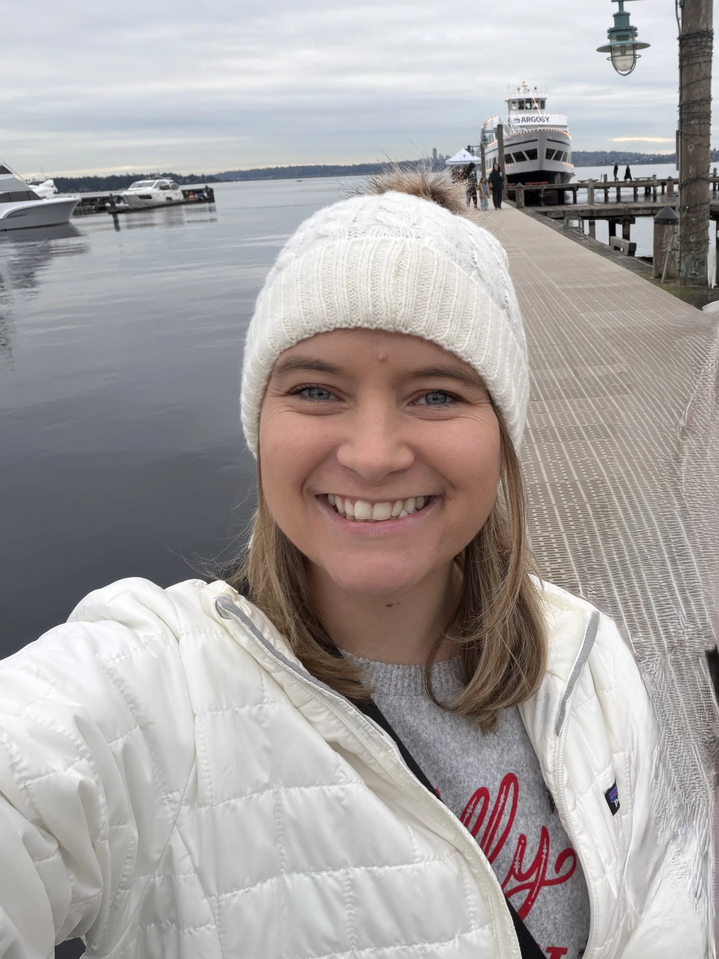 A smiling woman taking a selfie at a dock with boats and a large ferry in the background.