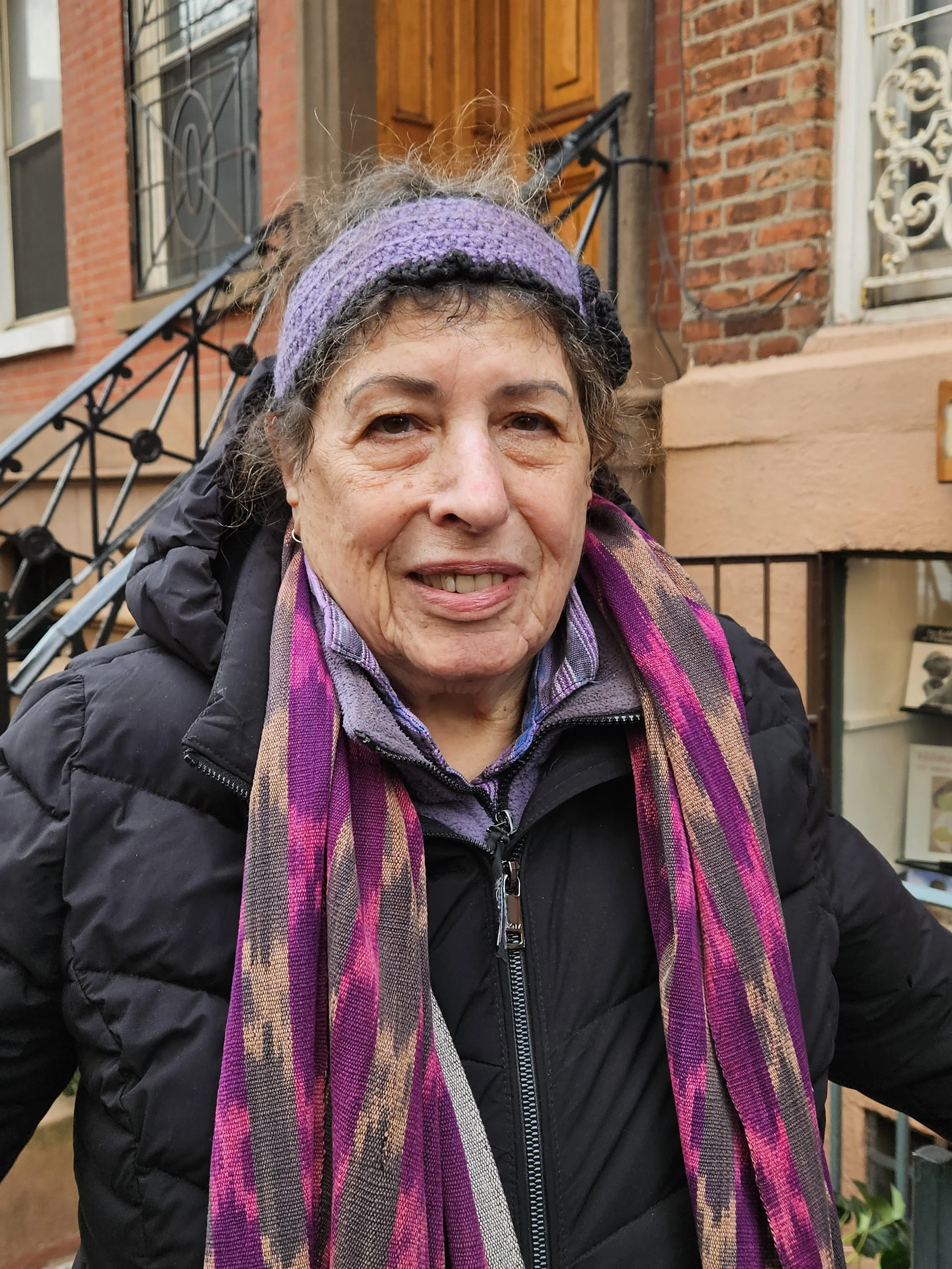 Close-up of Sylvia Mandel, wearing a blue cap outdoors with city buildings in the background.