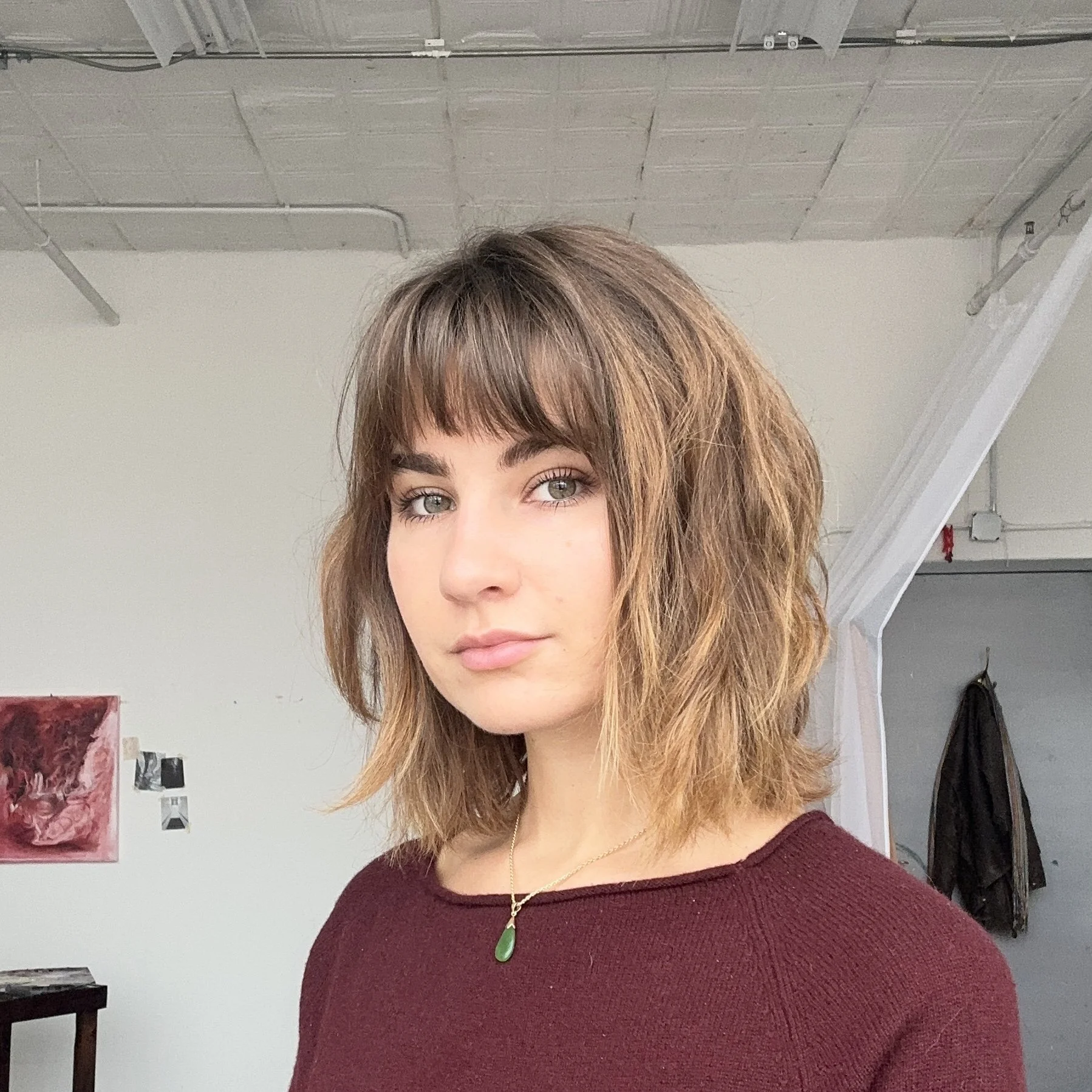 Close-up of Emily Fecsko, in an art studio. She is wearing a gray shirt and a beaded necklace. The background shows shelves with art supplies and a wooden art easel.