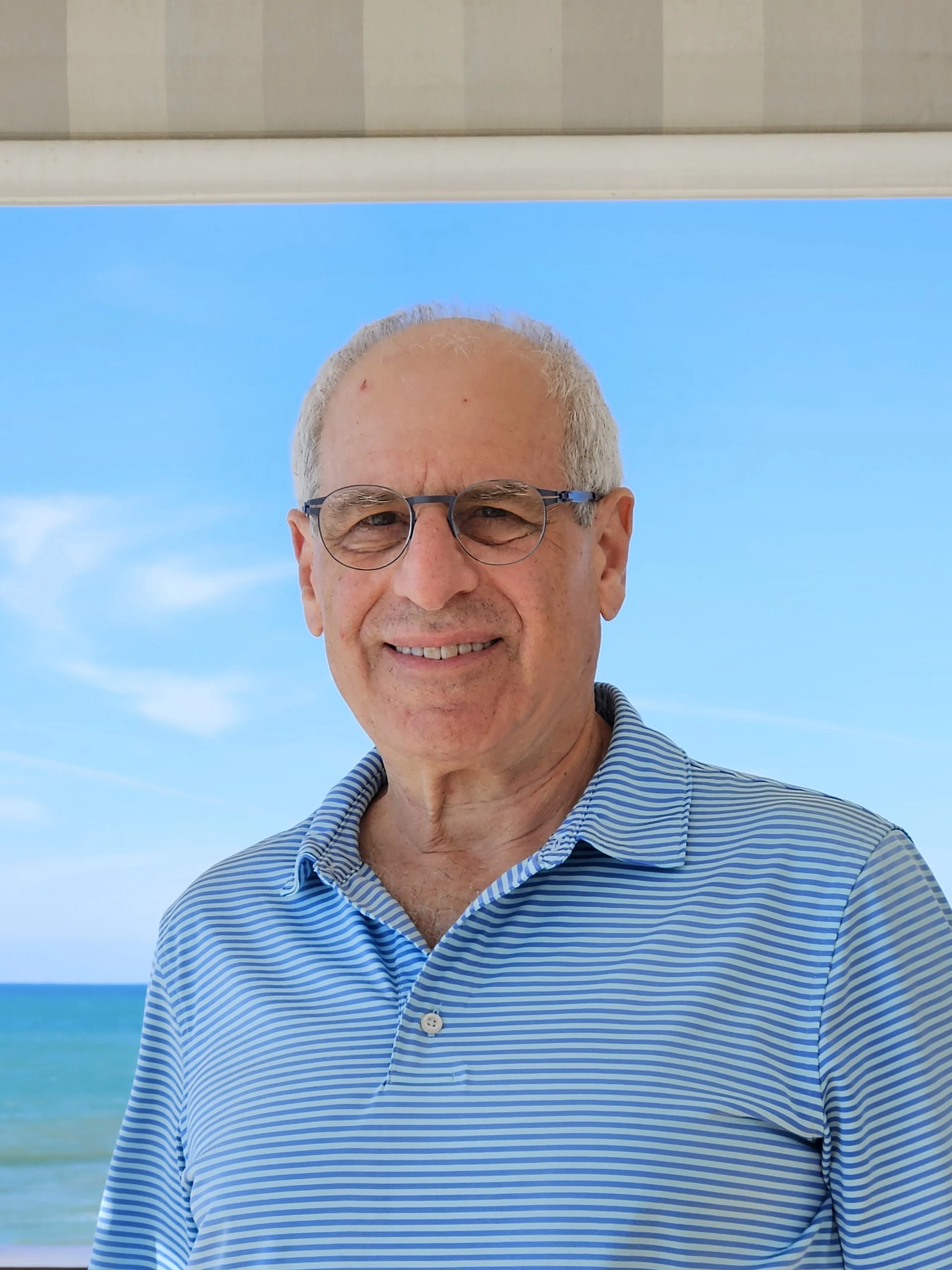 Close up of Dan Rabin, with glasses standing outdoors with a blue sky and ocean in the background.