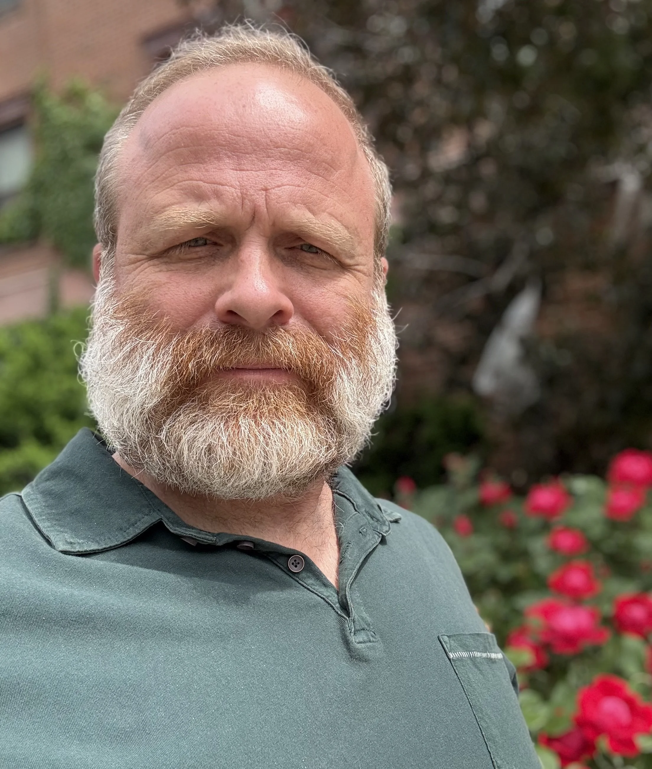 Close-up of Ryan Freeman, wearing a dark green collared shirt, outdoors with blurred flowers and greenery in the background.