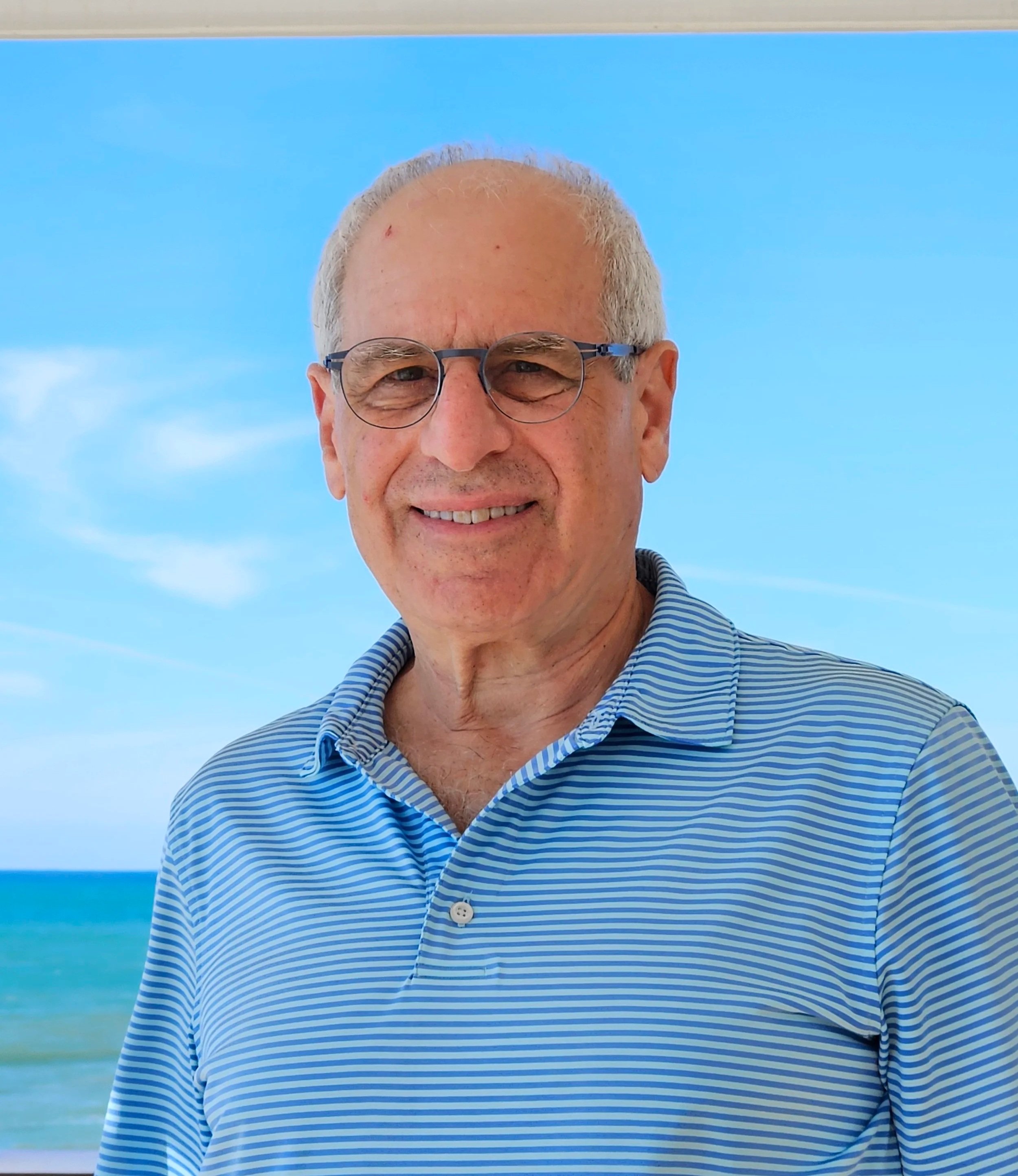 Close up of Dan Rabin, with glasses standing outdoors with a blue sky and ocean in the background.