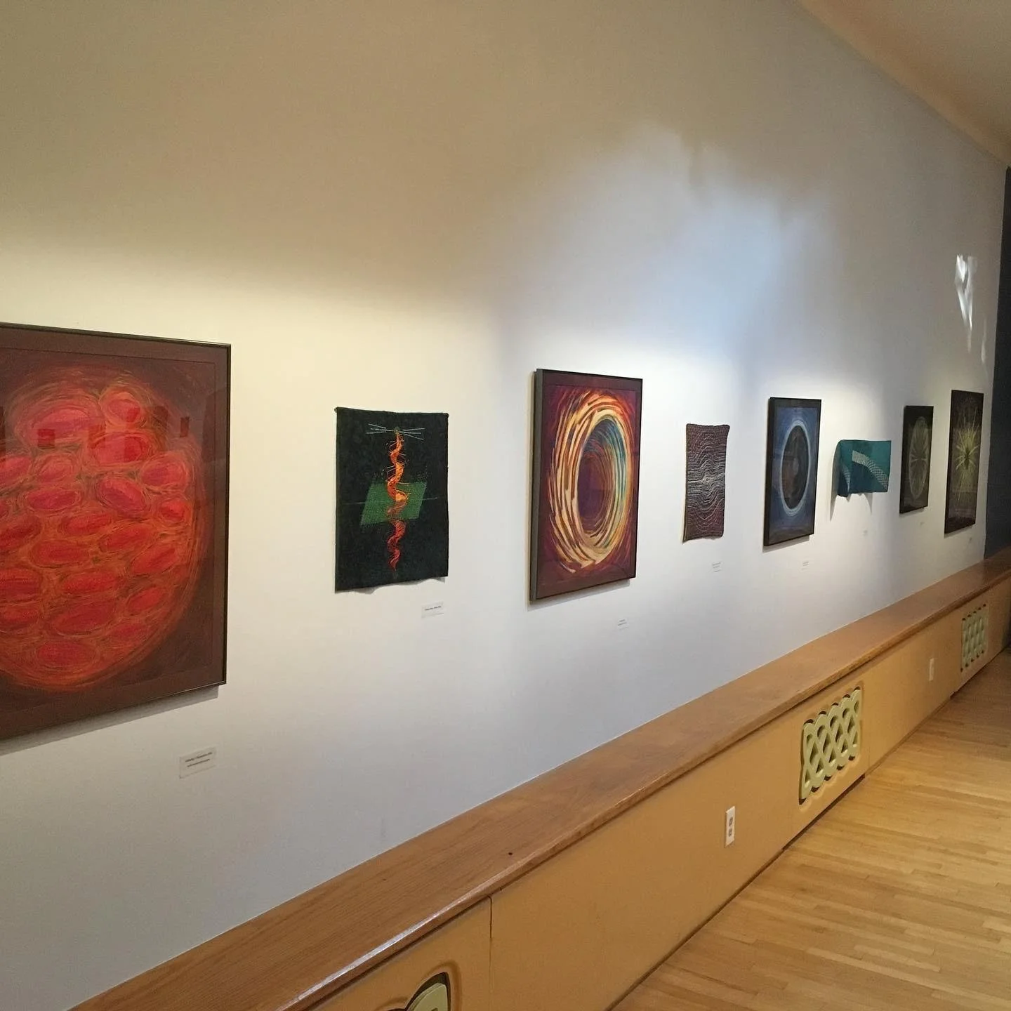 Art gallery wall displaying multiple abstract paintings with various colors and designs, illuminated by gallery lighting.