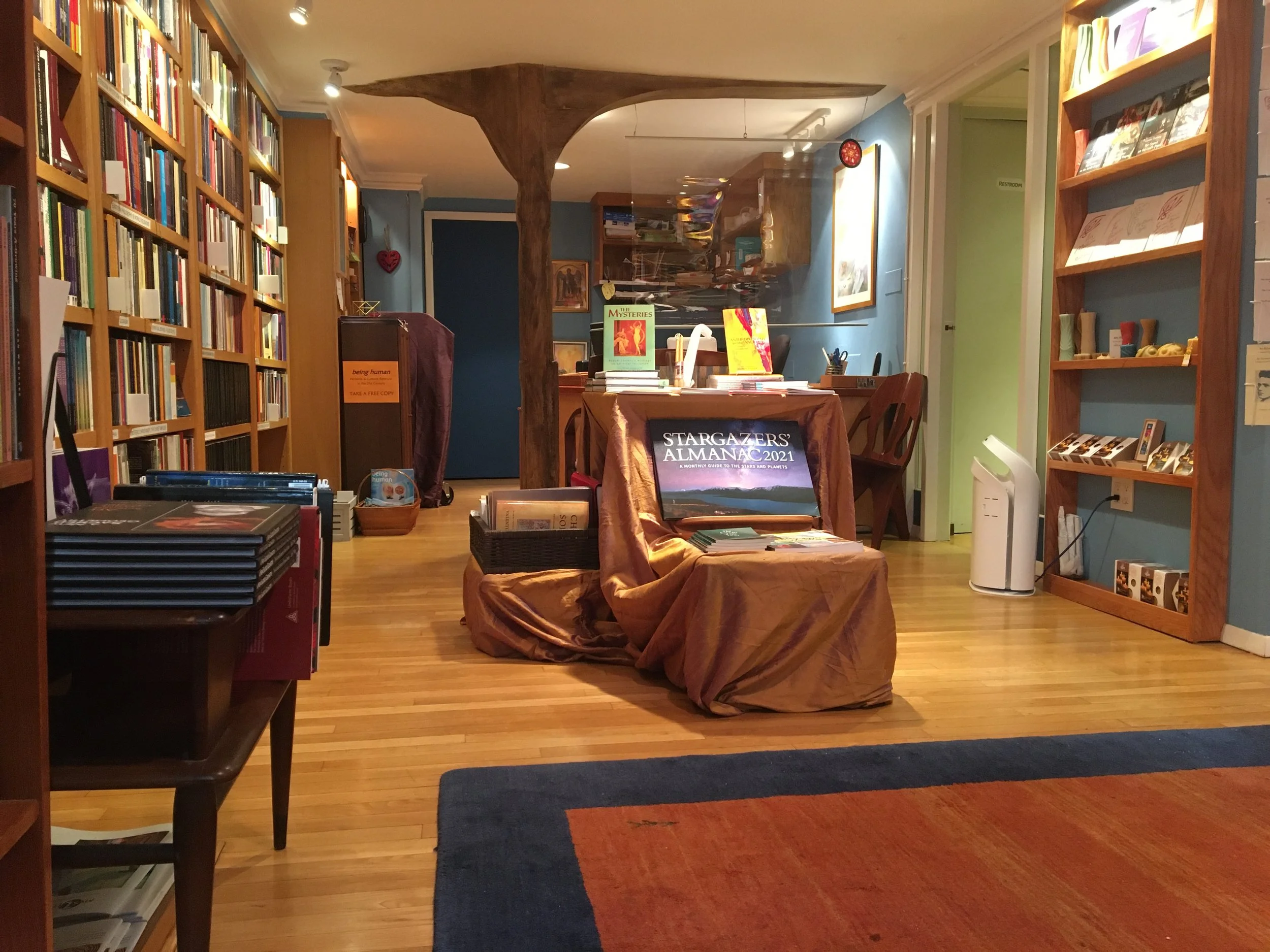 Interior of a bookstore or library with wooden floors, blue walls, and bookshelves filled with books. There are tables with displays, a blue door at the back, and a white air purifier on the right side.