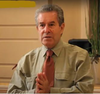 Middle-aged man with gray hair wearing a beige shirt and red tie, clasping his hands together, in an indoor setting.