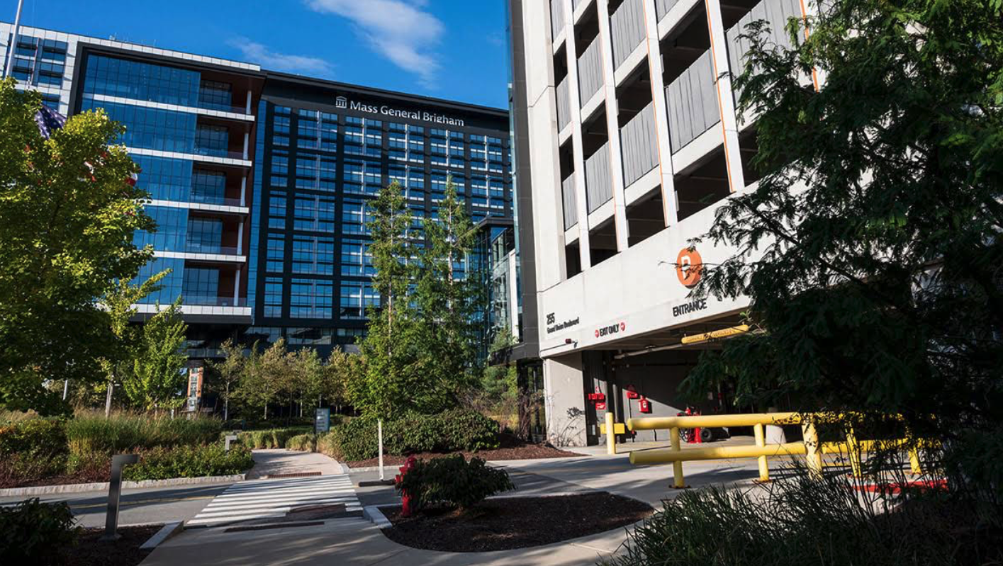 View of a city street with trees, bushes, a crosswalk, and modern buildings, including the Mass General Brigham hospital and a parking garage entrance with yellow barriers and exit signs.