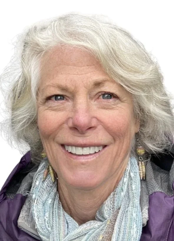 A smiling elderly woman with gray hair, wearing colorful earrings and a light-colored scarf, against a plain white background.