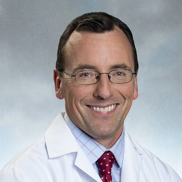 A smiling male doctor wearing glasses, a white lab coat, a light blue checked shirt, and a red tie, against a neutral background.