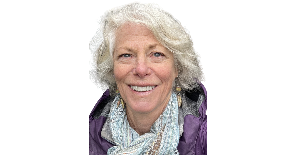 A smiling elderly woman with gray hair, wearing colorful earrings and a light-colored scarf, against a plain white background.