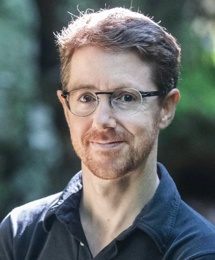 A man with glasses, short curly hair, and a beard smiling outdoors with greenery in the background.