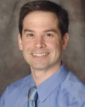 A man with dark hair, wearing a blue dress shirt and a light blue tie, smiling in front of a textured background.