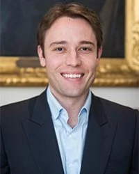 A young man with light brown hair wearing a dark blazer and light blue shirt, smiling in front of a dark background with a wooden frame.