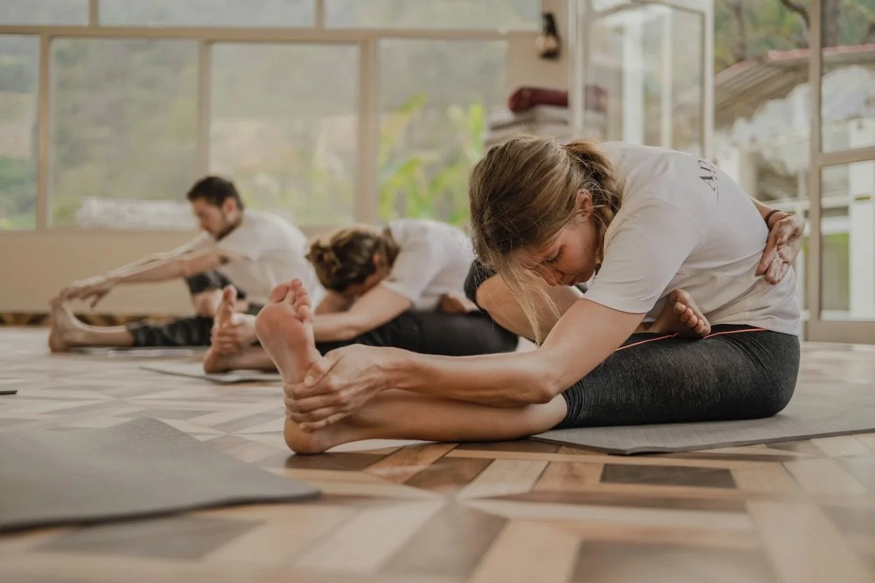 People participating in a yoga class stretching on mats in a bright room with large windows.