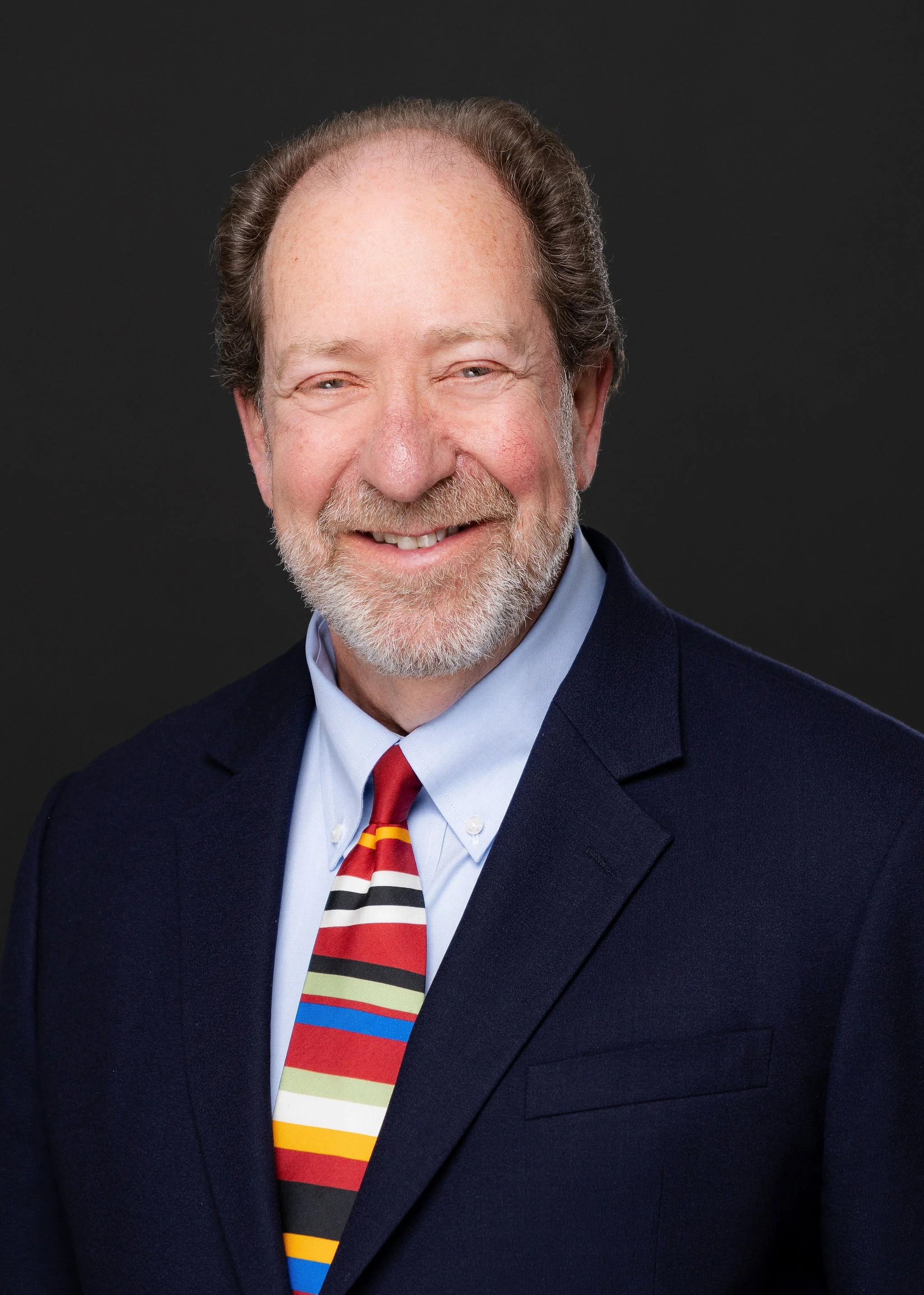 Portrait of a smiling older man with a beard, wearing a dark navy suit, light blue shirt, and a colorful striped tie, against a black background.
